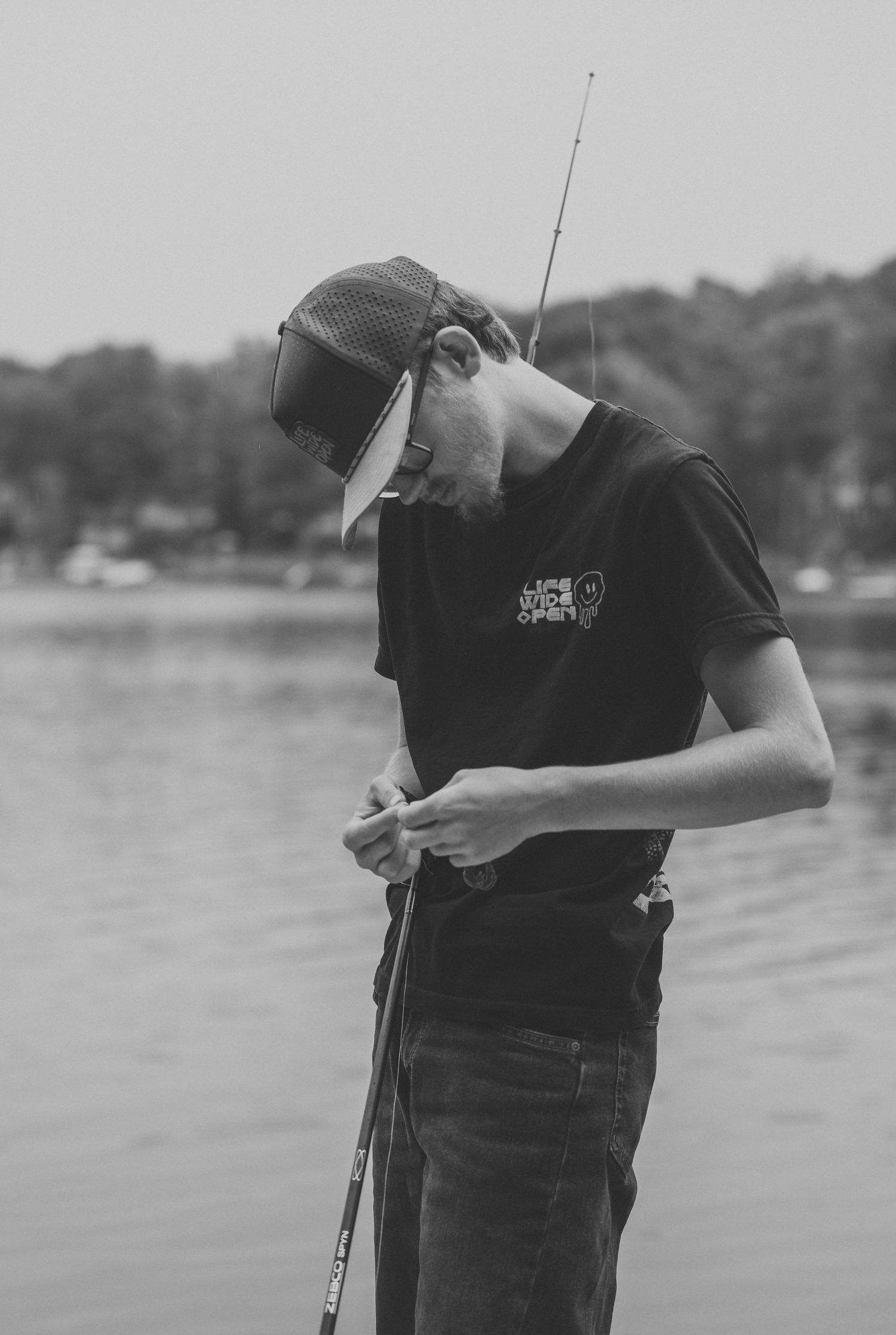 Black and white photo of a young man in a cap and graphic t-shirt tying his fishing line while holding a rod beside a calm lake.