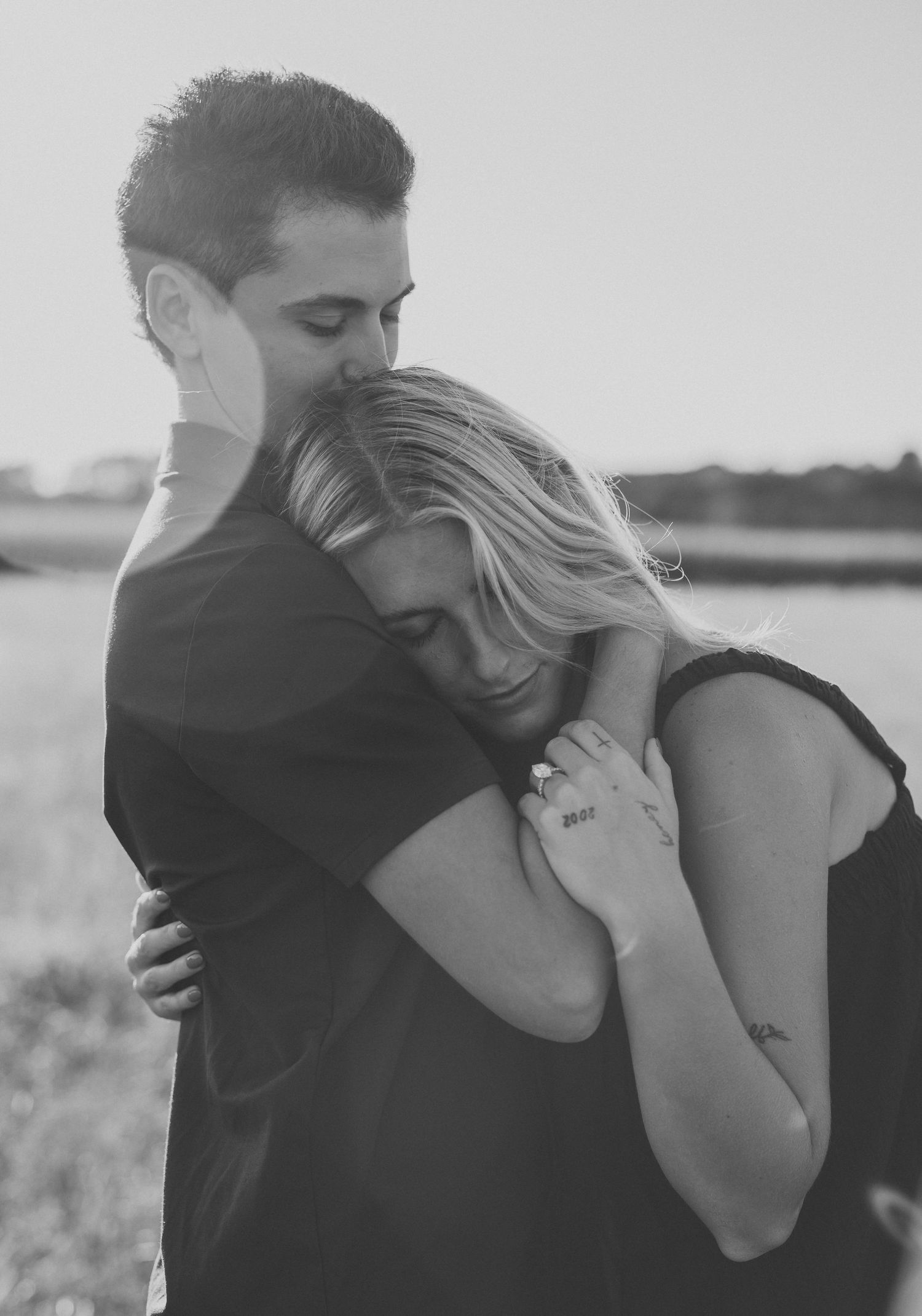 Black and white photo of a couple embracing in a grassy field, the woman resting her head on the man's chest as he kisses her forehead.