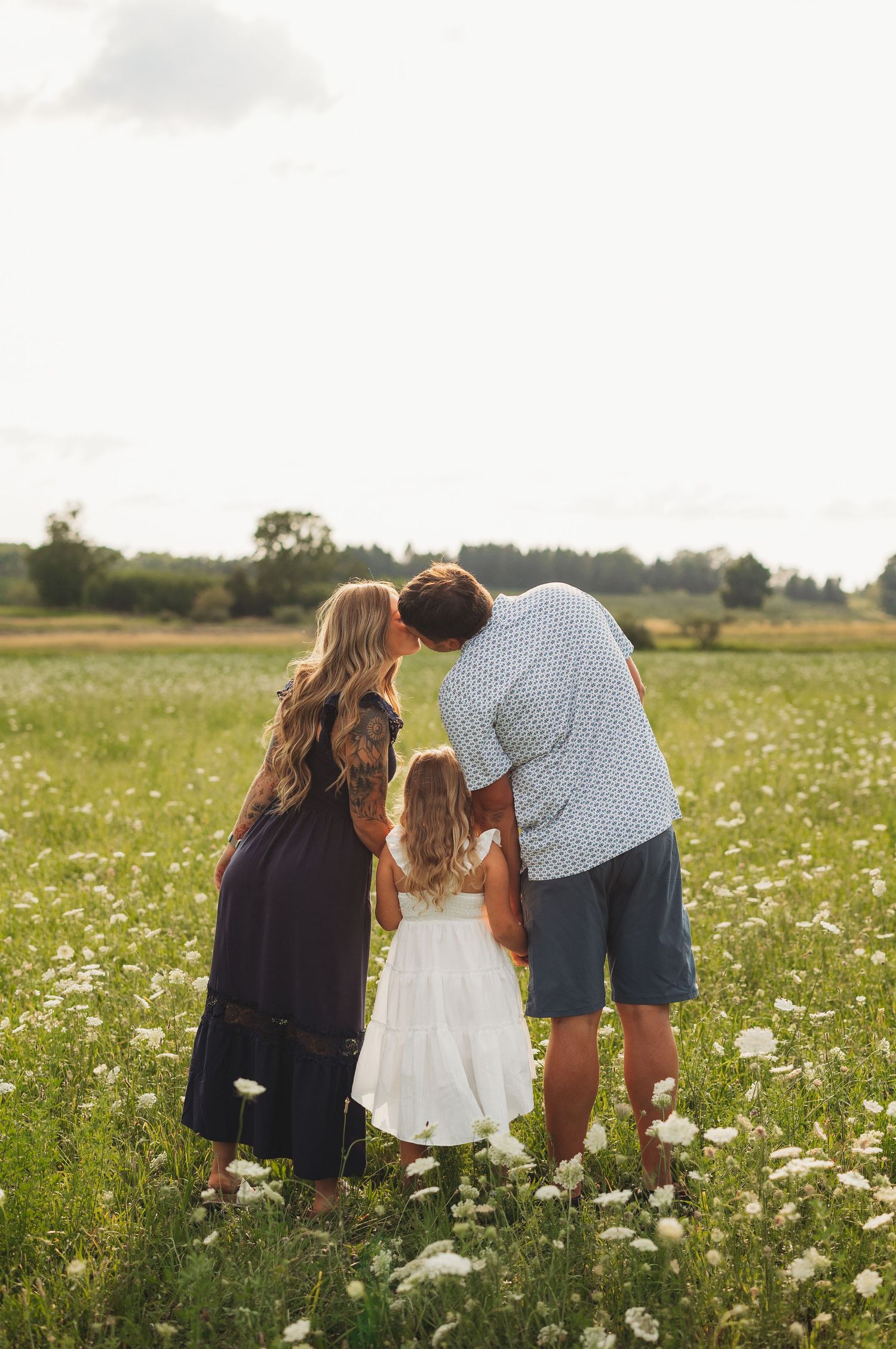 Family of three embracing in a sunny wildflower meadow, parents kissing as their young daughter in a white dress stands between them.