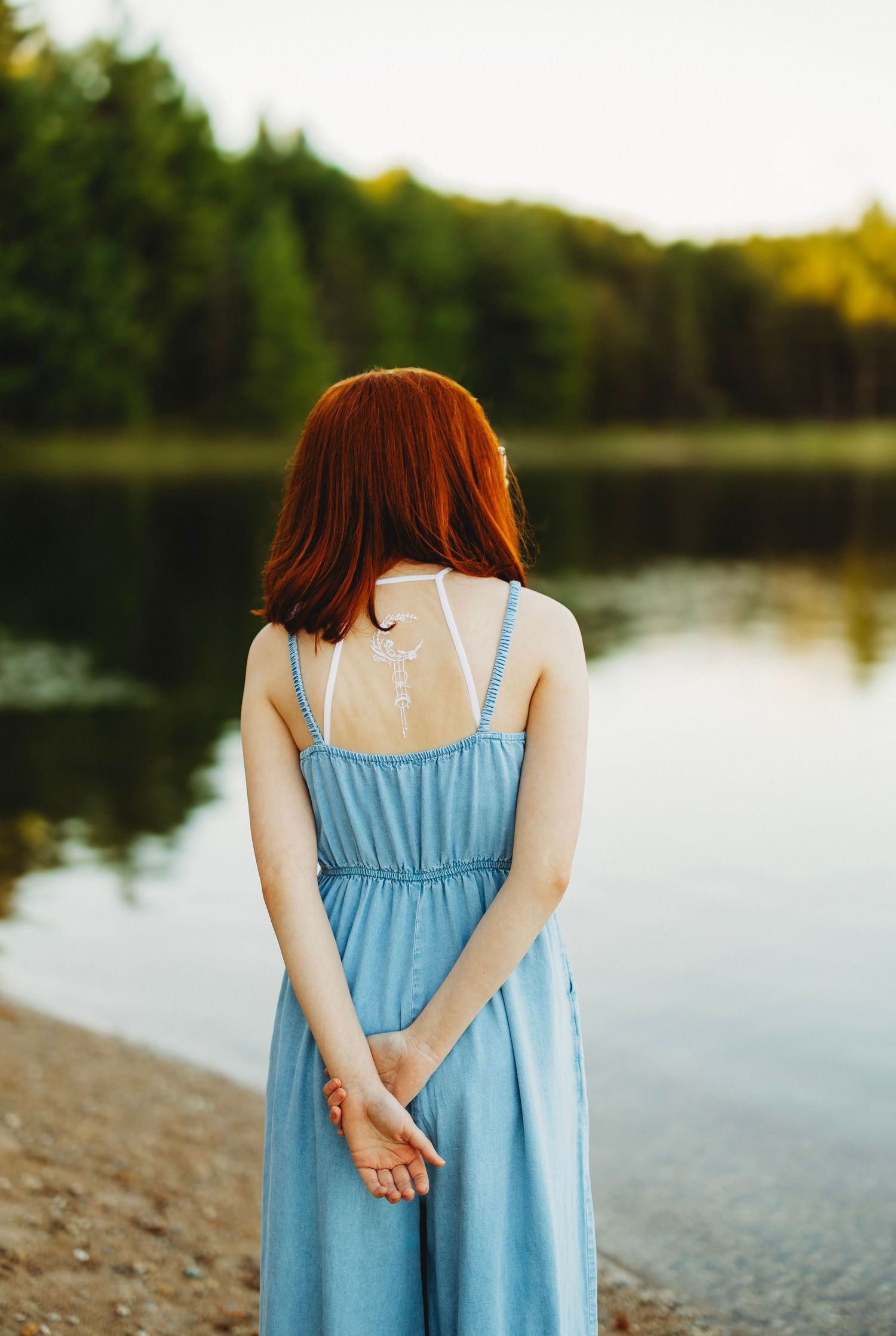 Woman with red hair in a blue denim dress stands with hands clasped behind her back, facing a calm lake and forest at sunset.