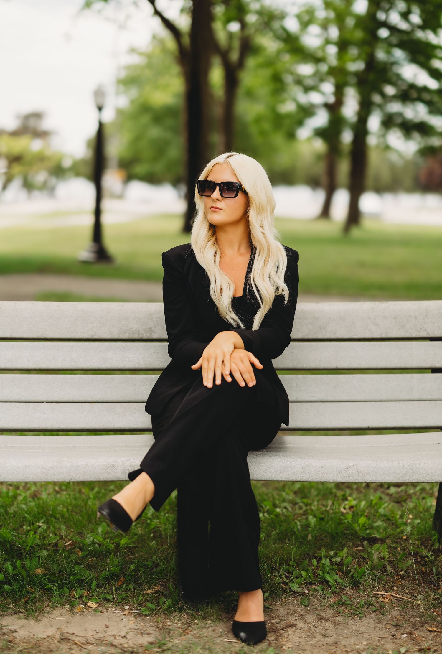 Blonde woman in black suit and sunglasses sitting on a white park bench surrounded by green trees.
