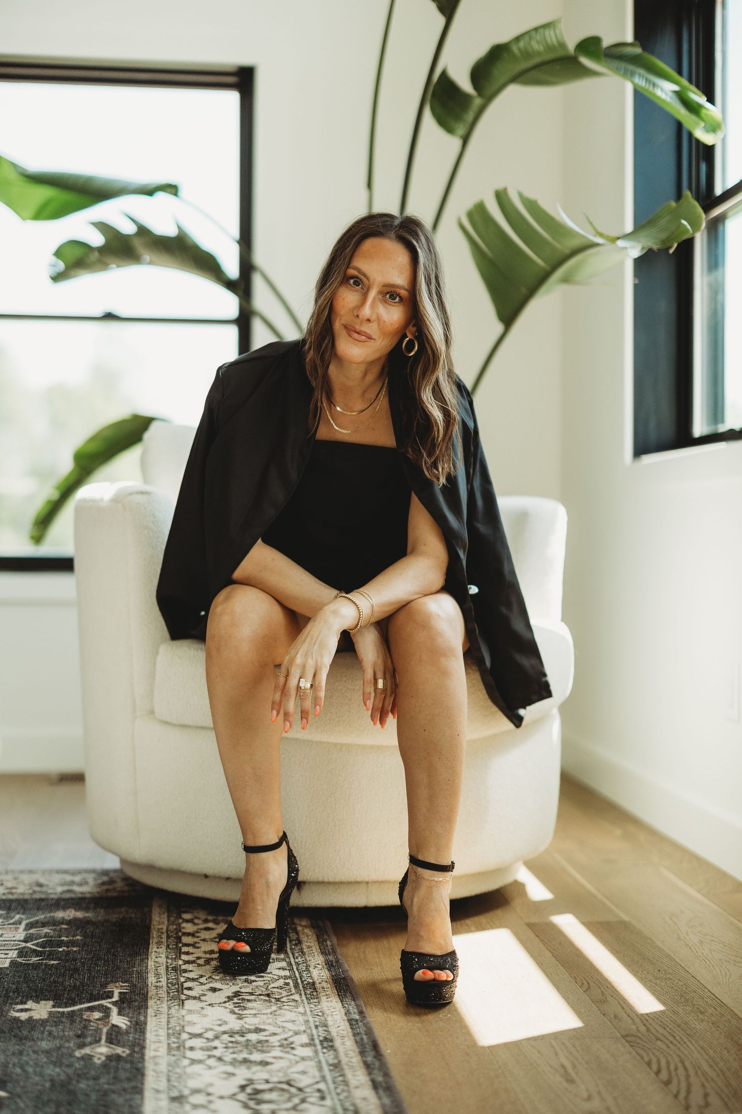 Woman in black blazer and heels sitting in white armchair with tropical plant backdrop in bright modern room.