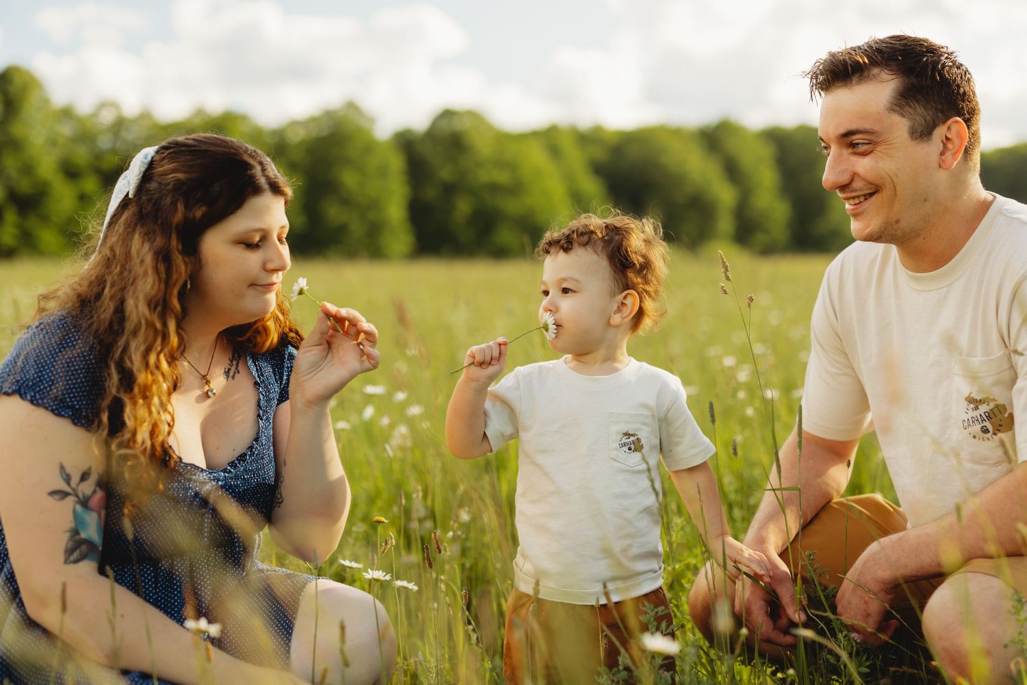 A young family sitting in a sunny meadow with wildflowers, as a toddler smells a flower while parents smile warmly.