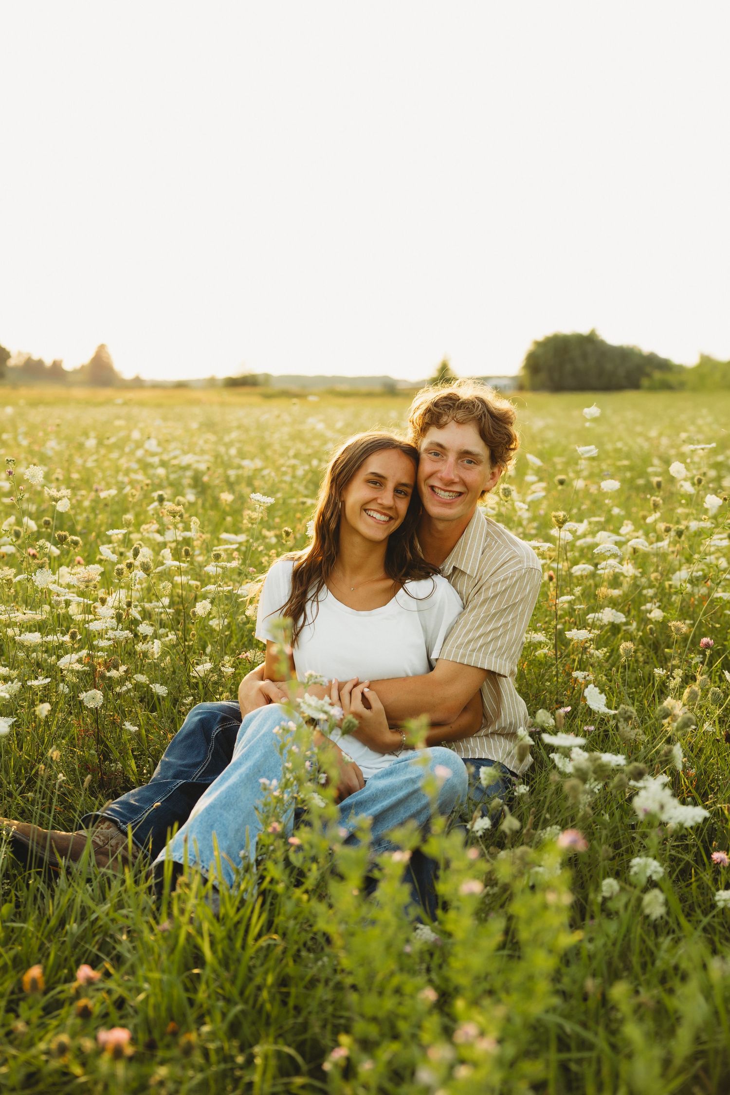 A couple sits embracing in a sunlit wildflower meadow, smiling warmly at the camera during golden hour.