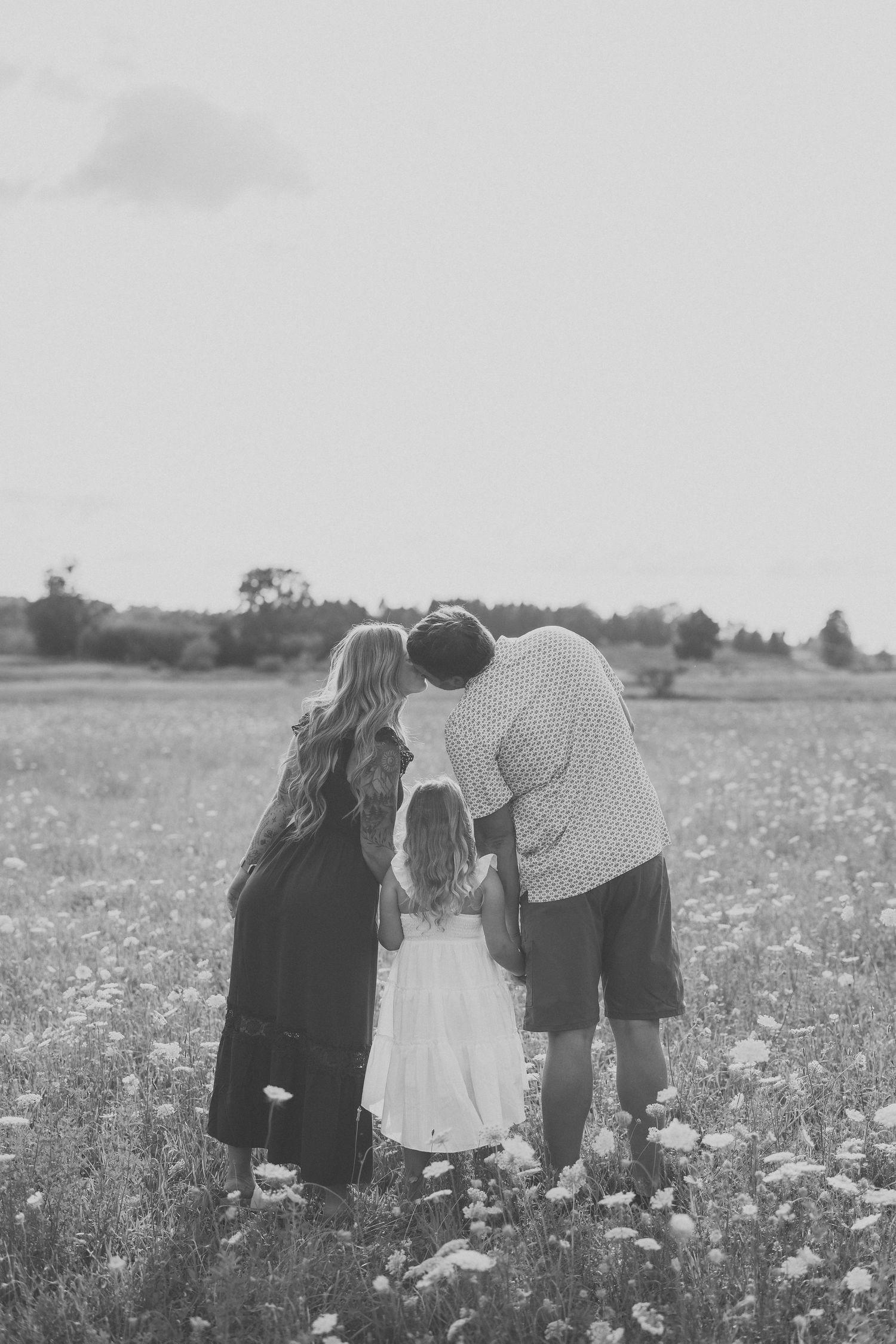 Black and white photo of a family of three standing in a wildflower field, parents kissing over their young daughter.