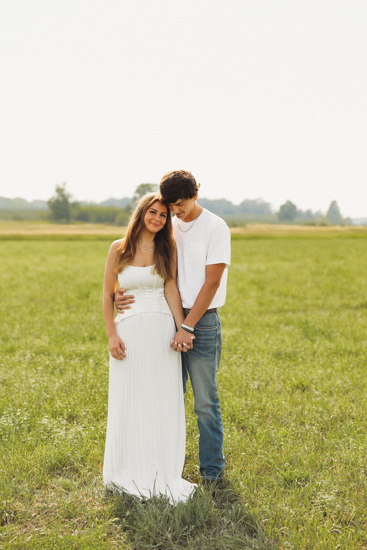 Couple standing in green field, woman in white dress with man in white tee and jeans, embracing tenderly.