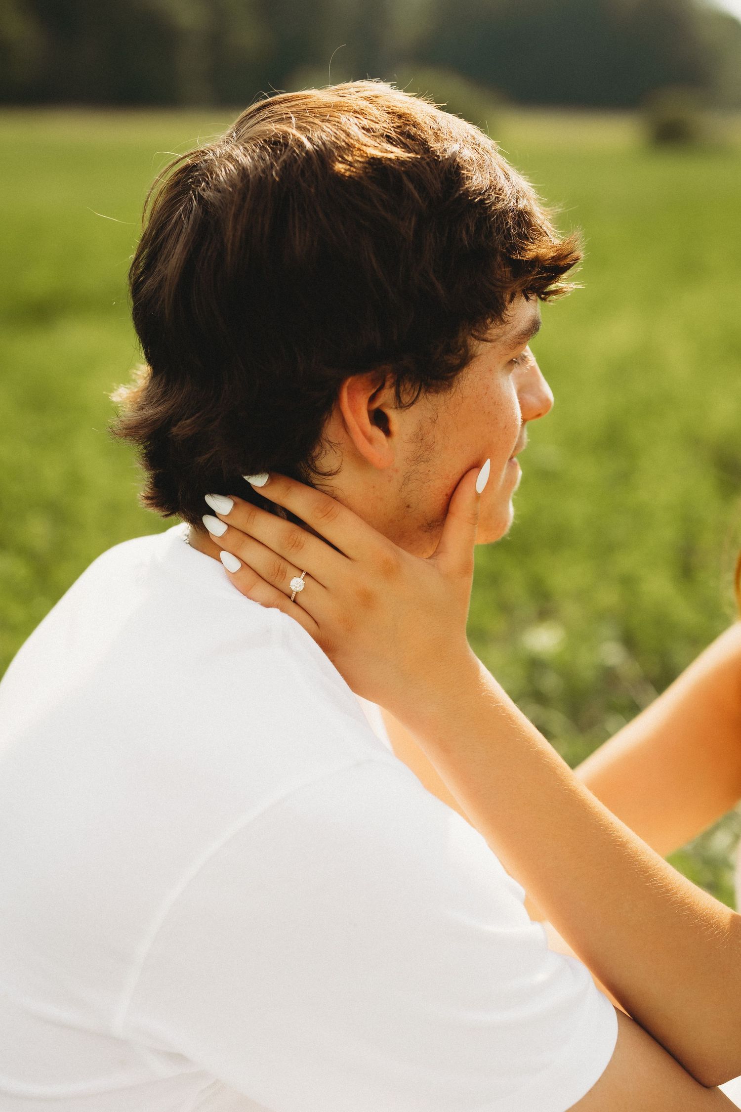 A woman with a diamond ring tenderly holds a man's face in a sunny green field.