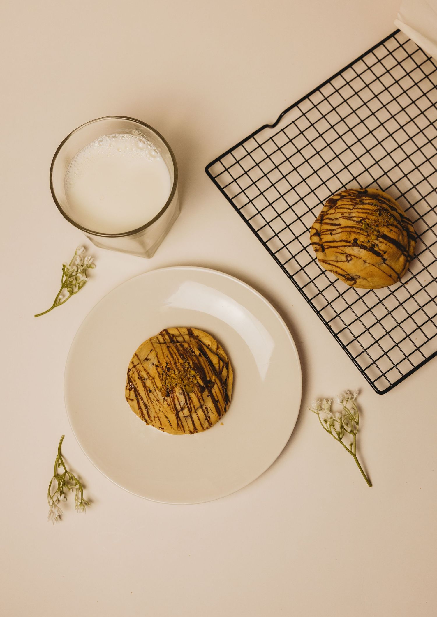 Cookie with chocolate drizzle on white plate beside glass of milk and cooling rack with small white flowers.