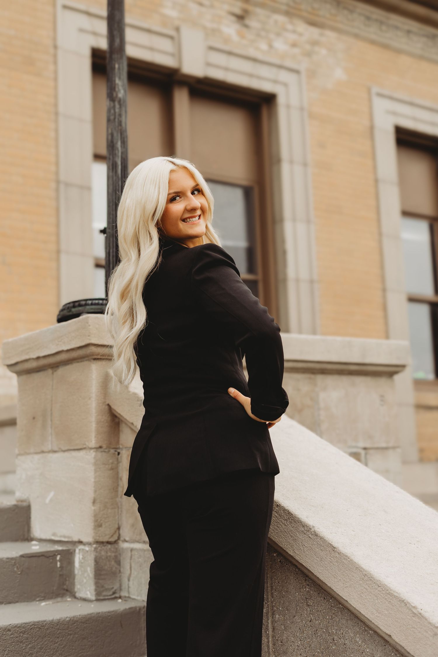 A smiling woman with long blonde hair wearing a black suit poses on stone steps outside a brick building.
