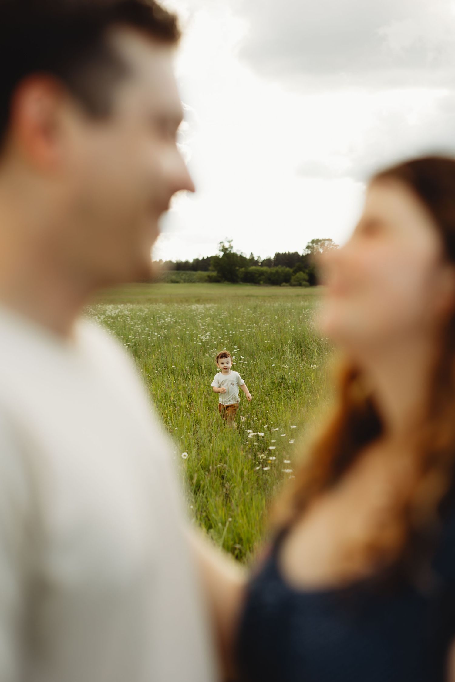 A young child runs through a green meadow while his parents stand in the foreground in soft focus.