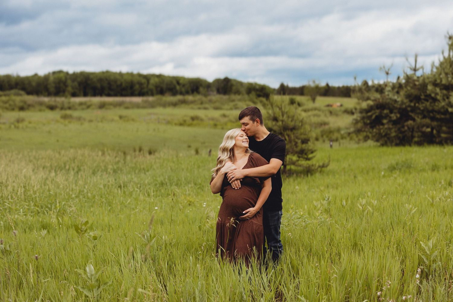 A couple embraces in a lush green field, the woman visibly pregnant, surrounded by trees under a cloudy sky.