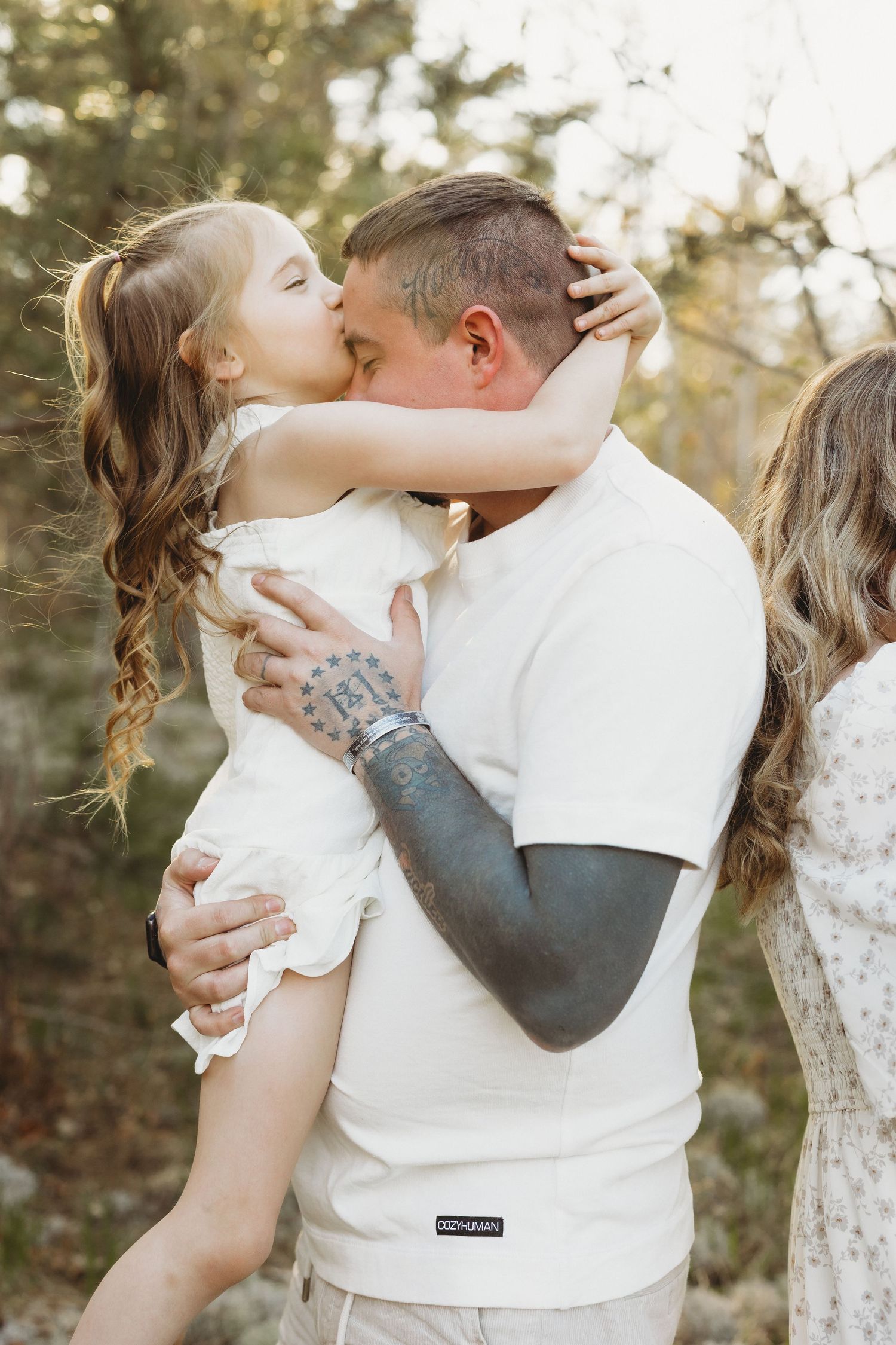 A young girl kisses a tattooed man in a white shirt while he holds her outdoors.