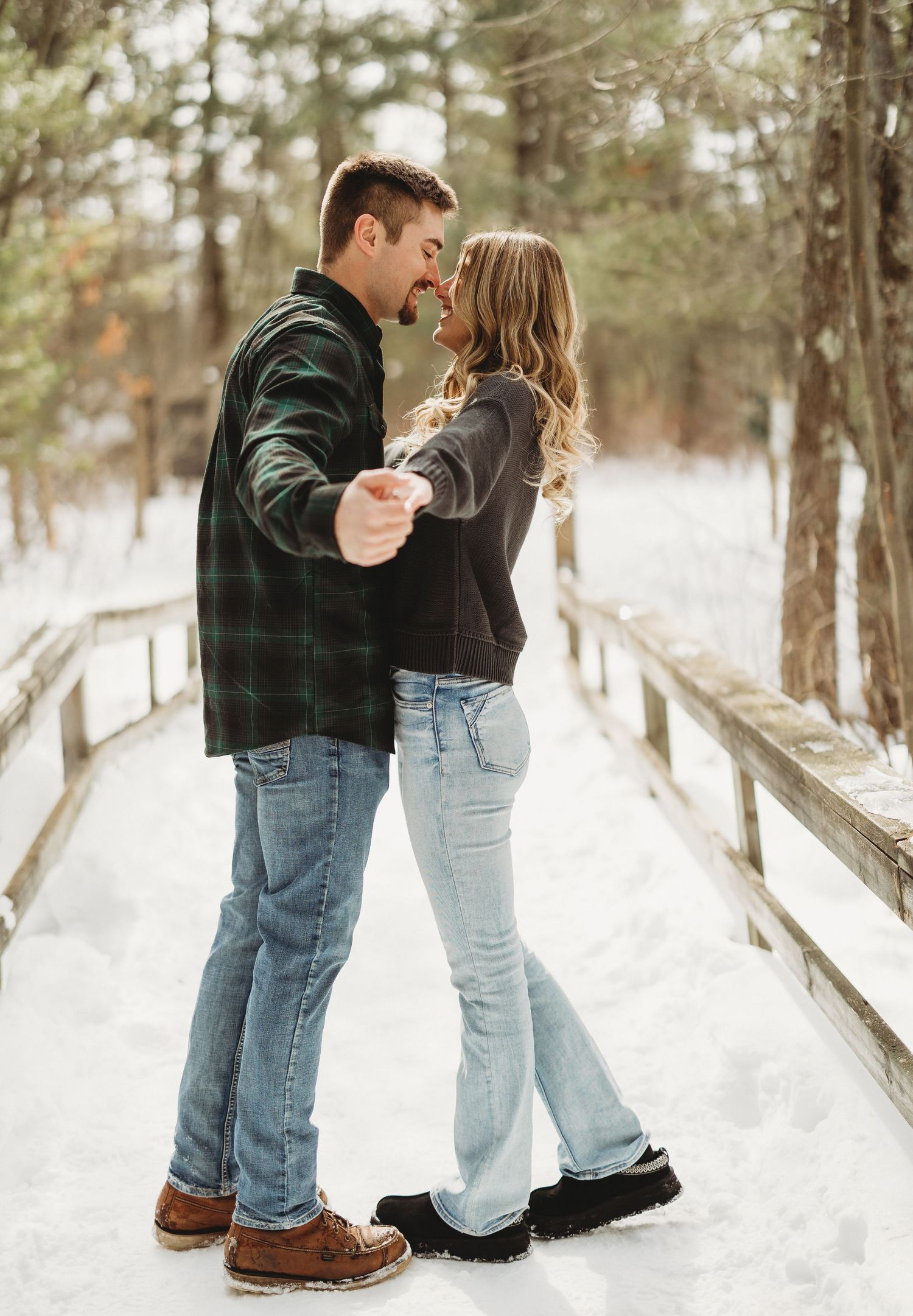 Playful couple photos in the snow with pine trees and a wooden fence in background.