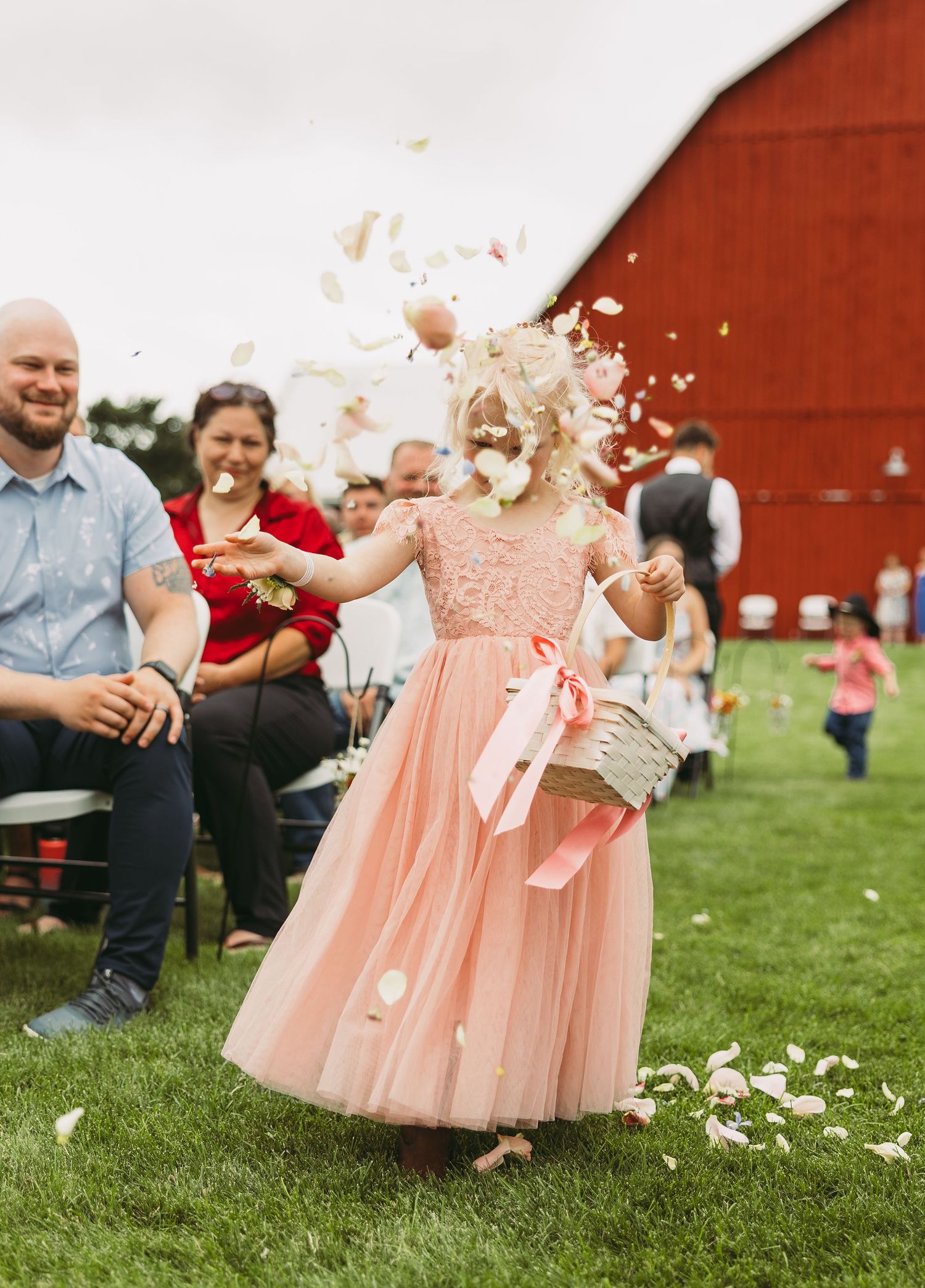 Young flower girl in pink dress throwing petals in front of a red barn.