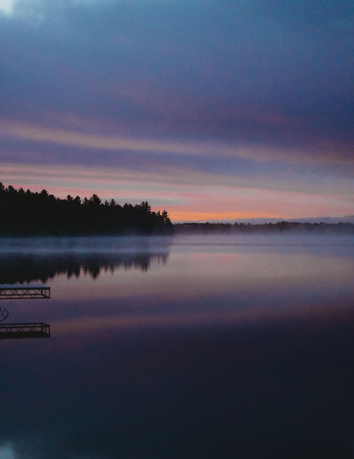 Panoramic view of a tranquil lake at sunset with silhouetted trees and moody purple clouds reflecting on calm waters.