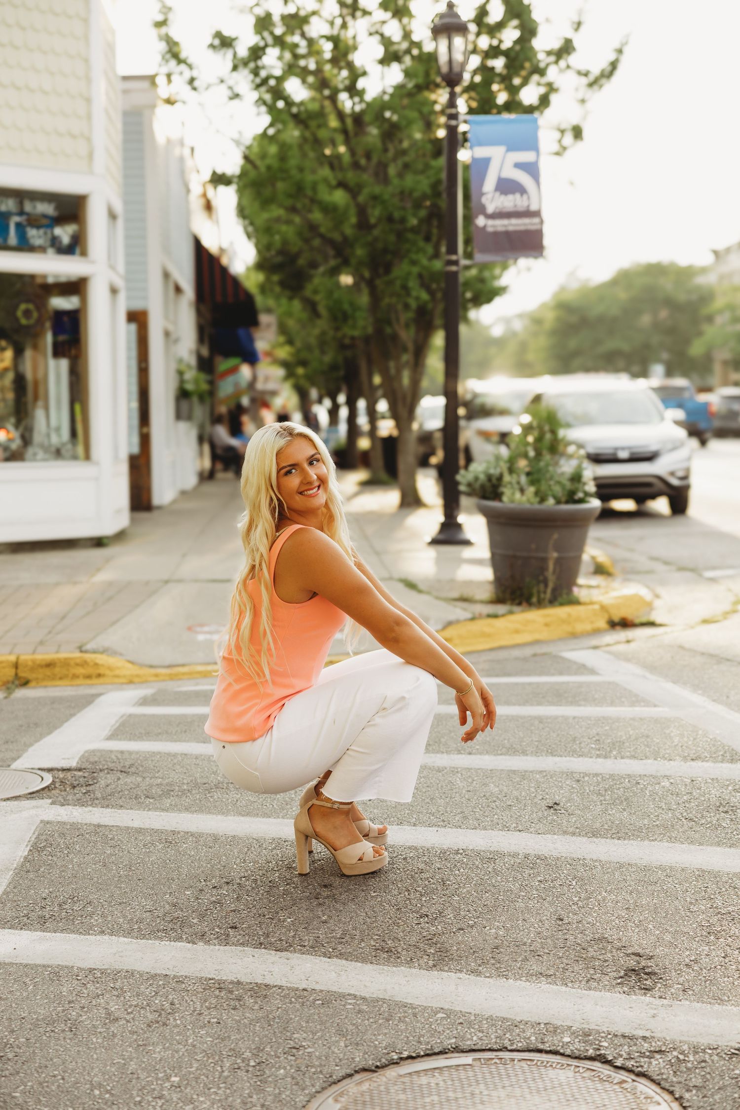Smiling woman with long blonde hair crouches on a crosswalk in a coral top, white pants and platform heels on a sunny small-town street.