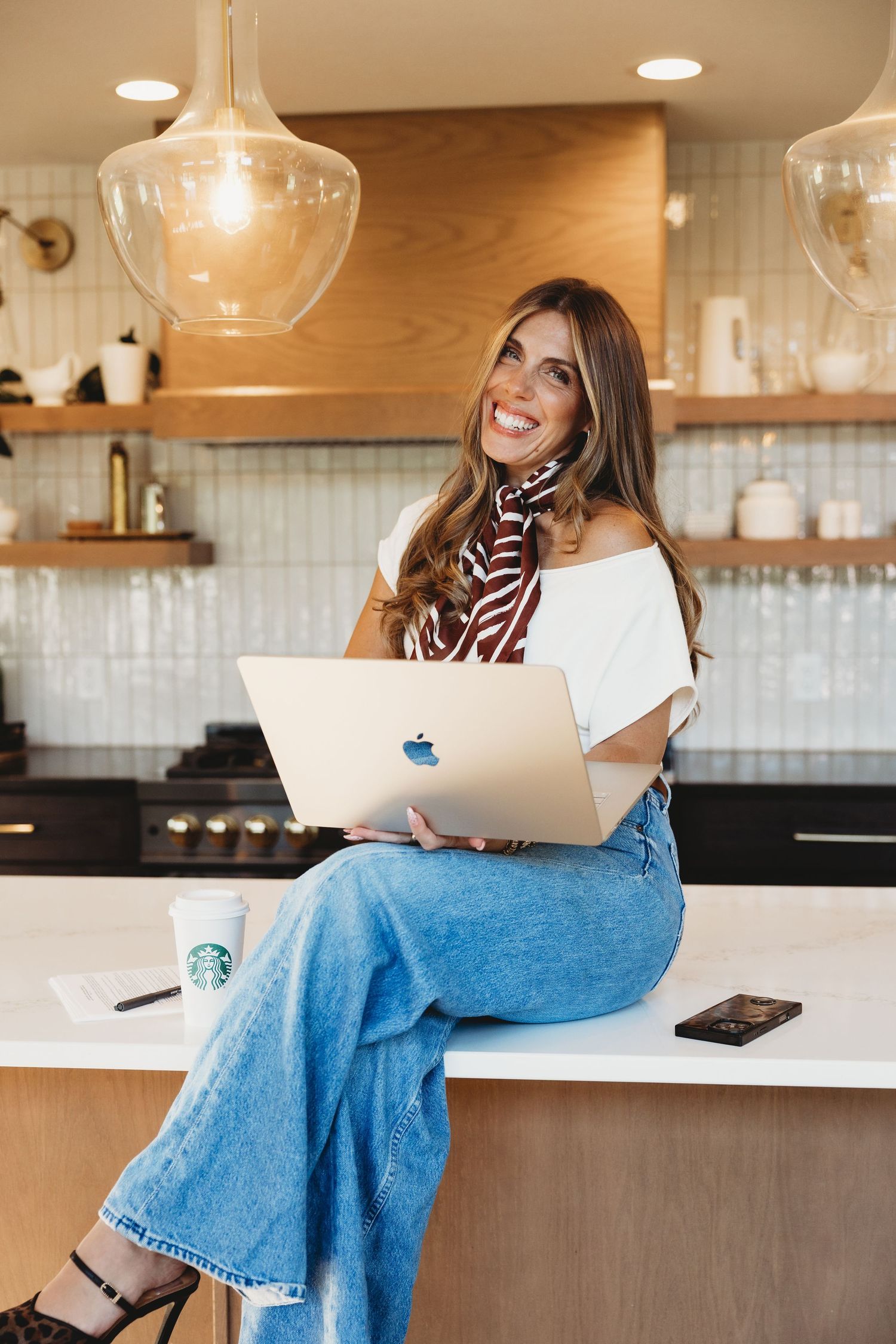 Smiling woman in wide-leg jeans and a striped scarf sits on a kitchen counter working on a laptop beside a Starbucks coffee cup.