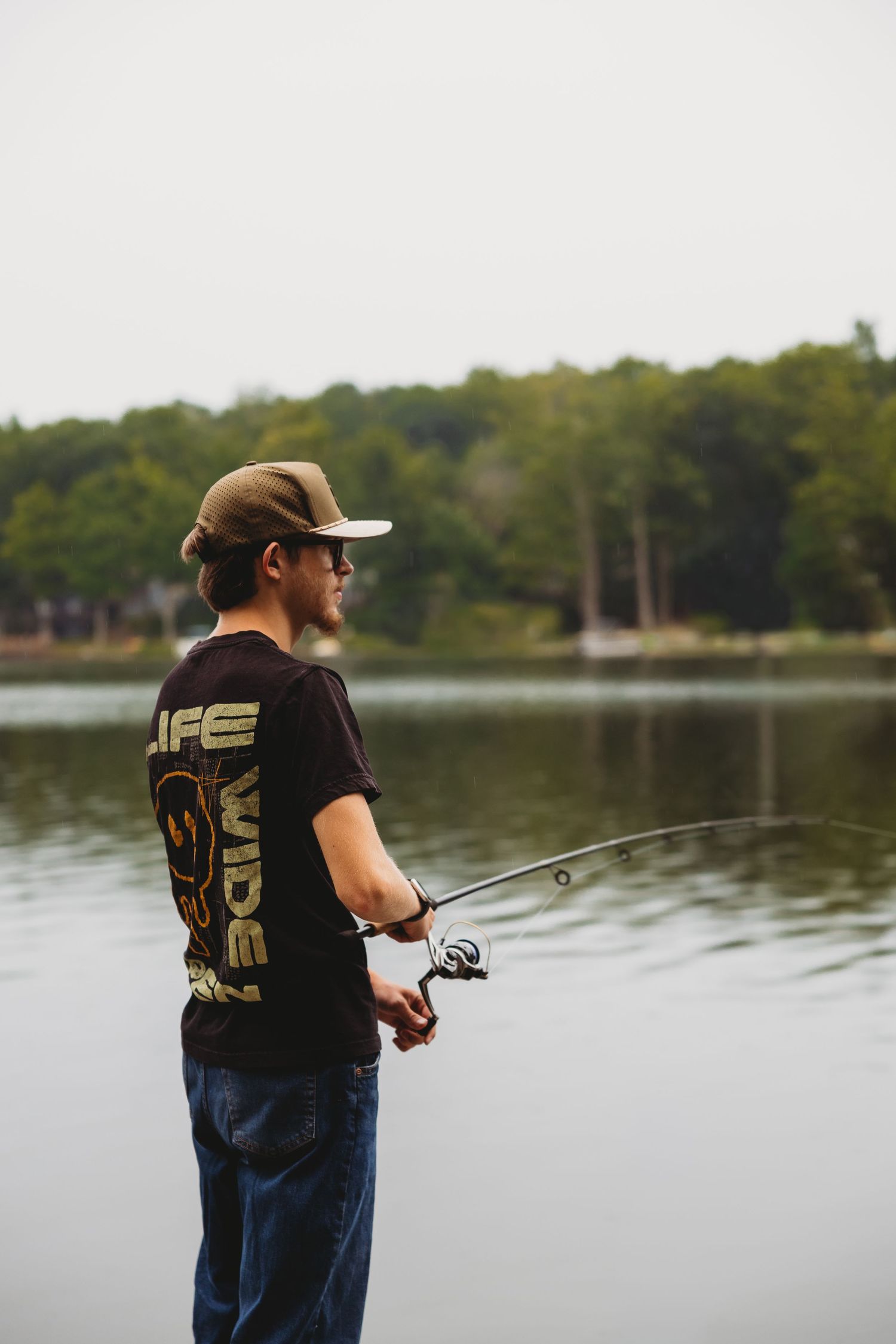 Man in a cap, sunglasses and graphic t-shirt fishing with a spinning rod from the shore of a calm lake with trees in the background.