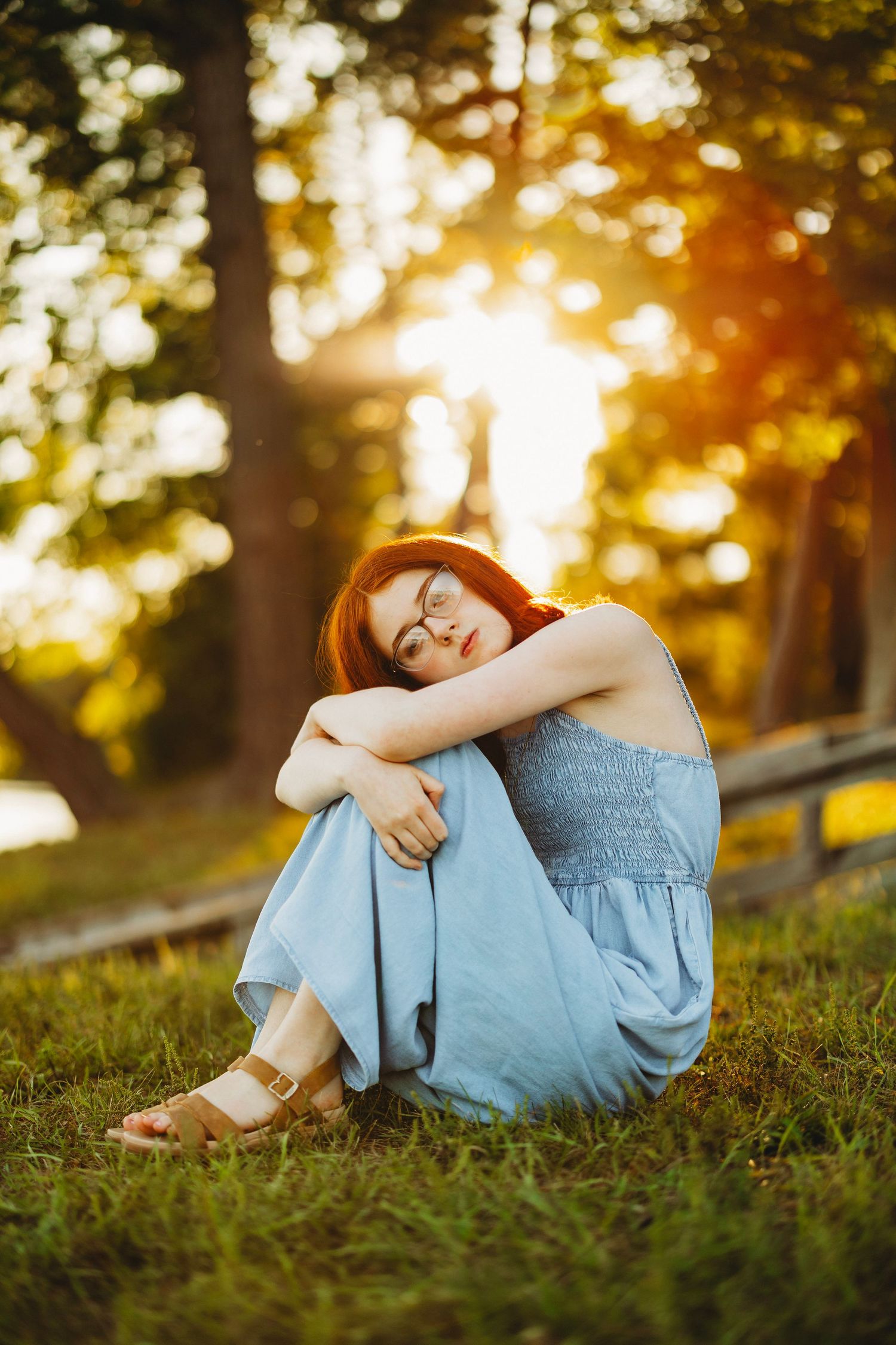 Red-haired woman in glasses and a light blue sundress sits hugging her knees on a grassy field in warm golden sunset light.