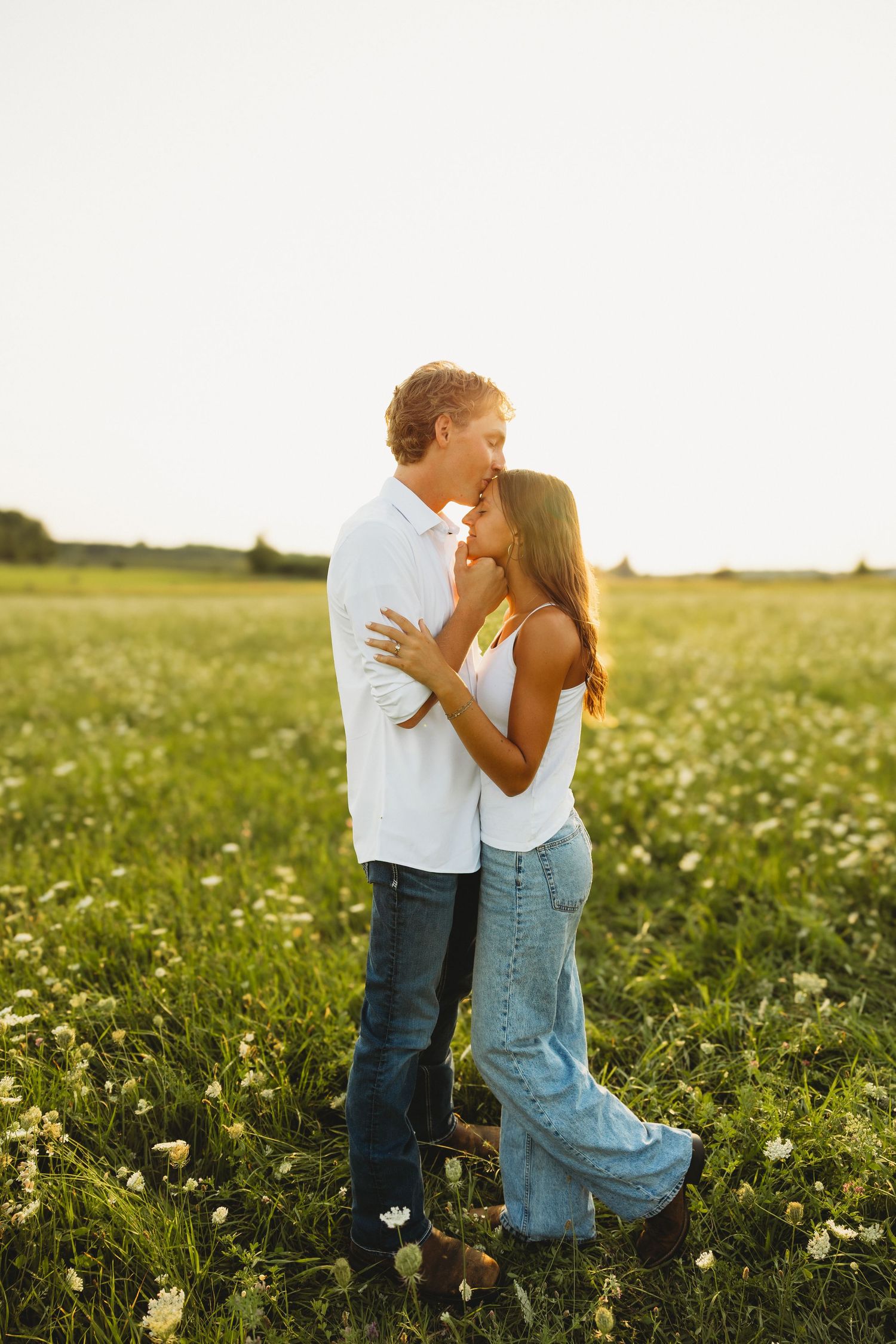 Couple embracing and almost kissing while standing in a blooming green field at sunset.