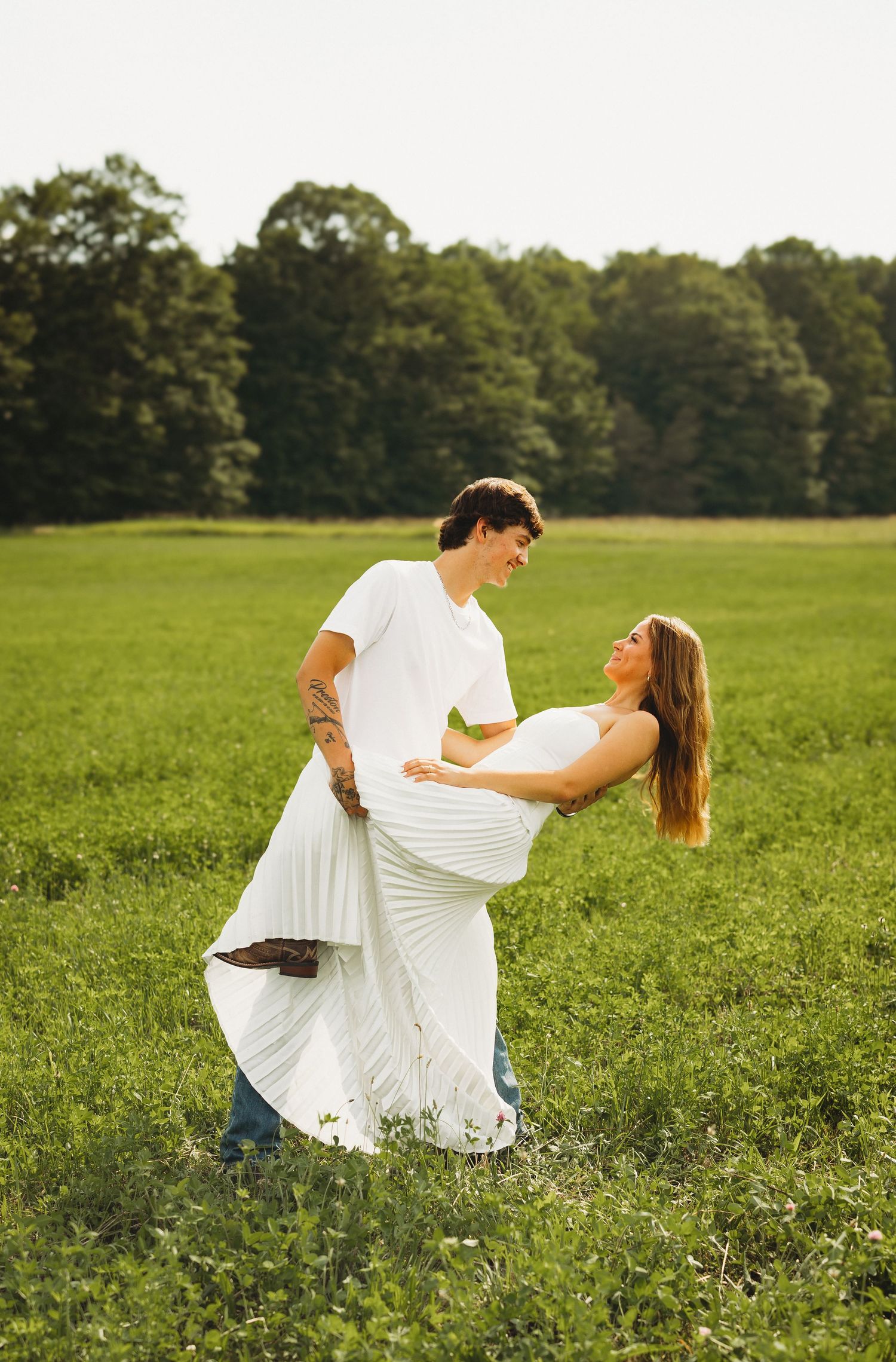 Couple dancing in a green field, woman in white dress dips back joyfully, trees in background.