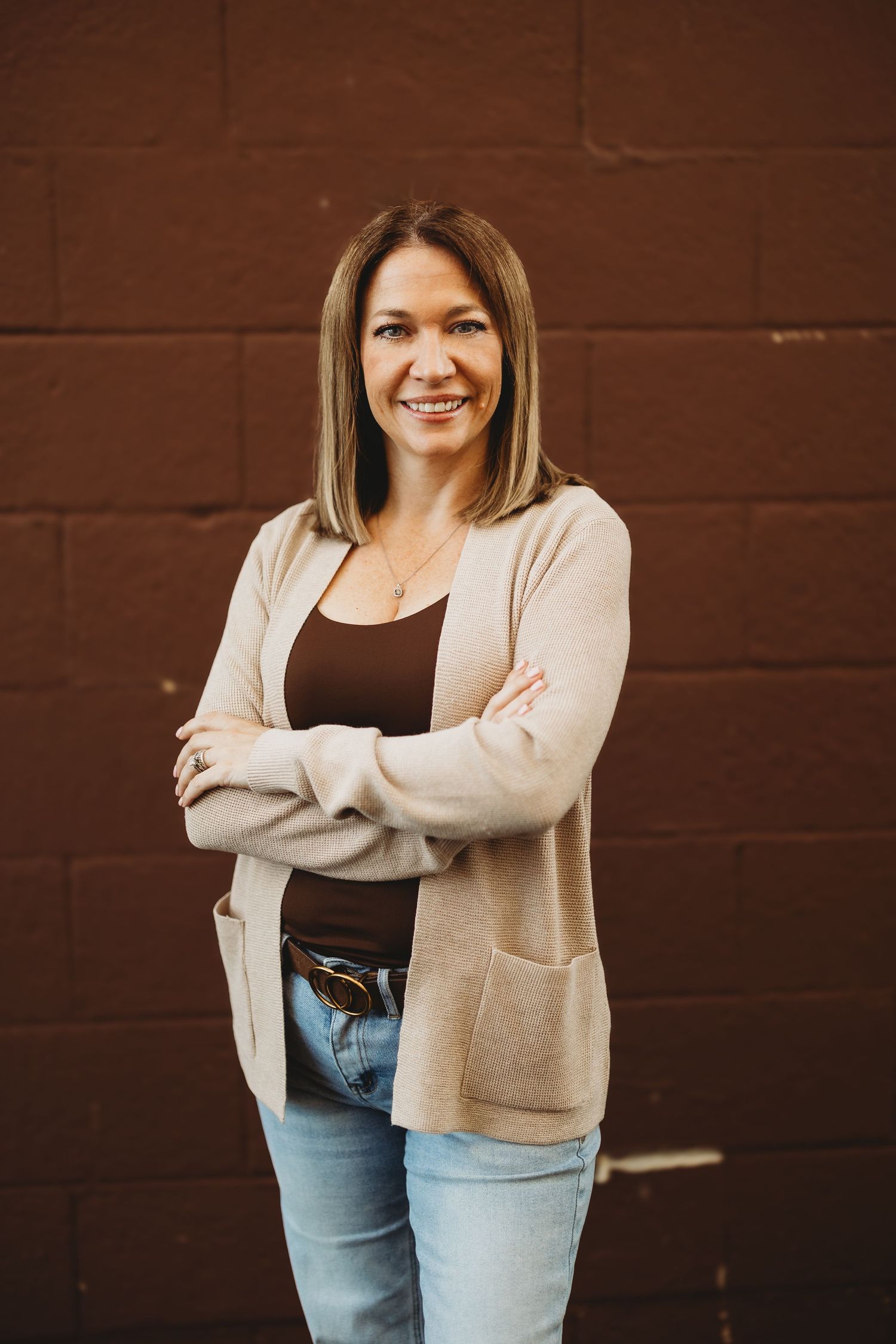 Smiling woman with crossed arms in beige cardigan and jeans posing against a dark brown brick wall.