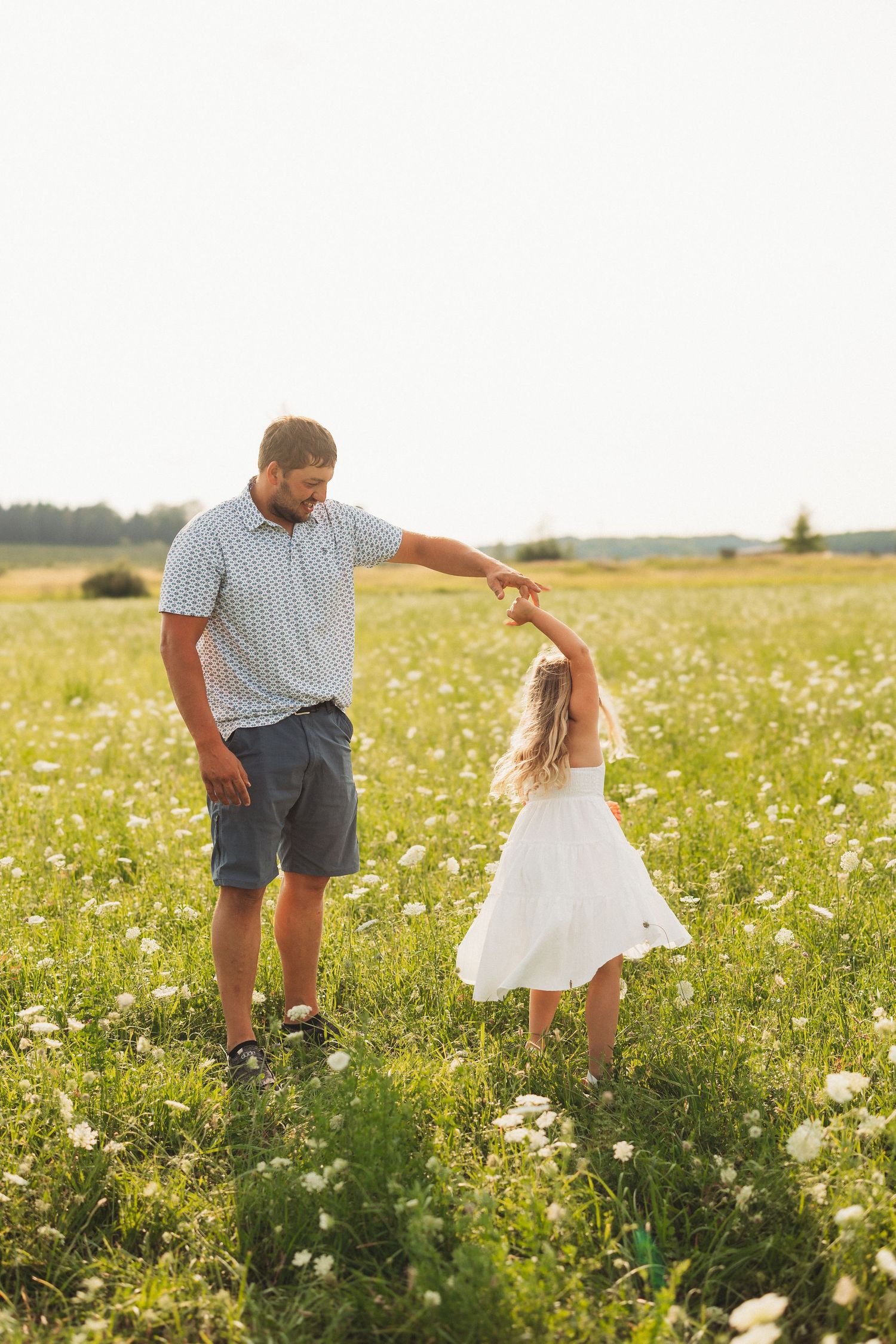 Father twirling his young daughter in a white dress in a sunny green meadow filled with wildflowers.