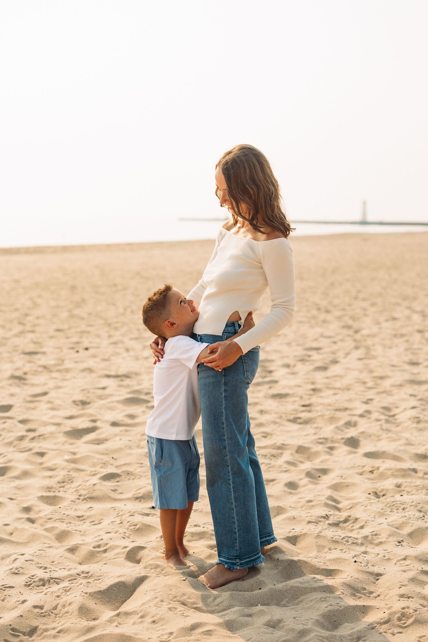 A young boy hugging his pregnant mother's belly on a sandy beach, both wearing casual white tops and jeans.