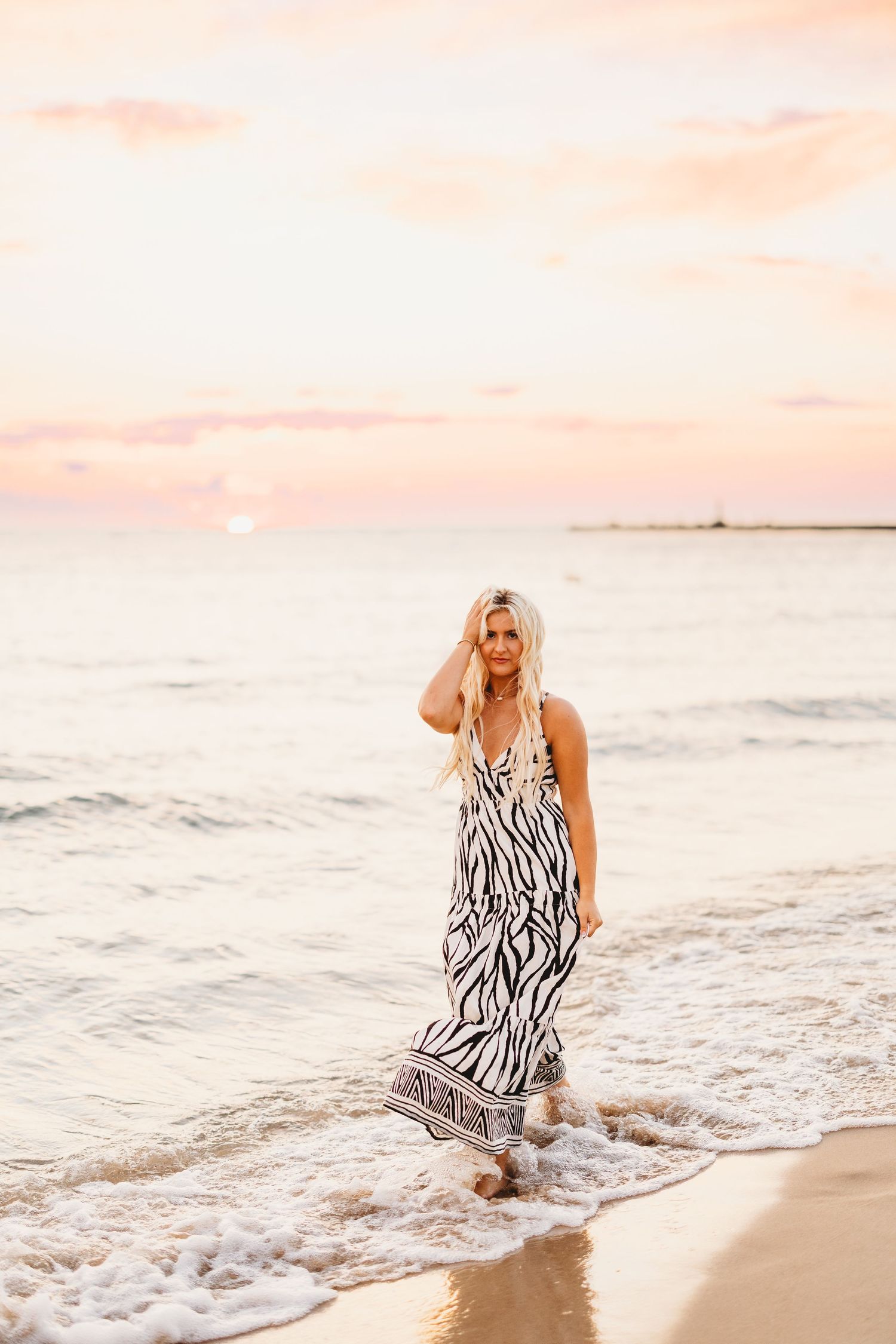 Woman in a black and white zebra print maxi dress walks along the shoreline as gentle waves roll in under a pink sunset sky.