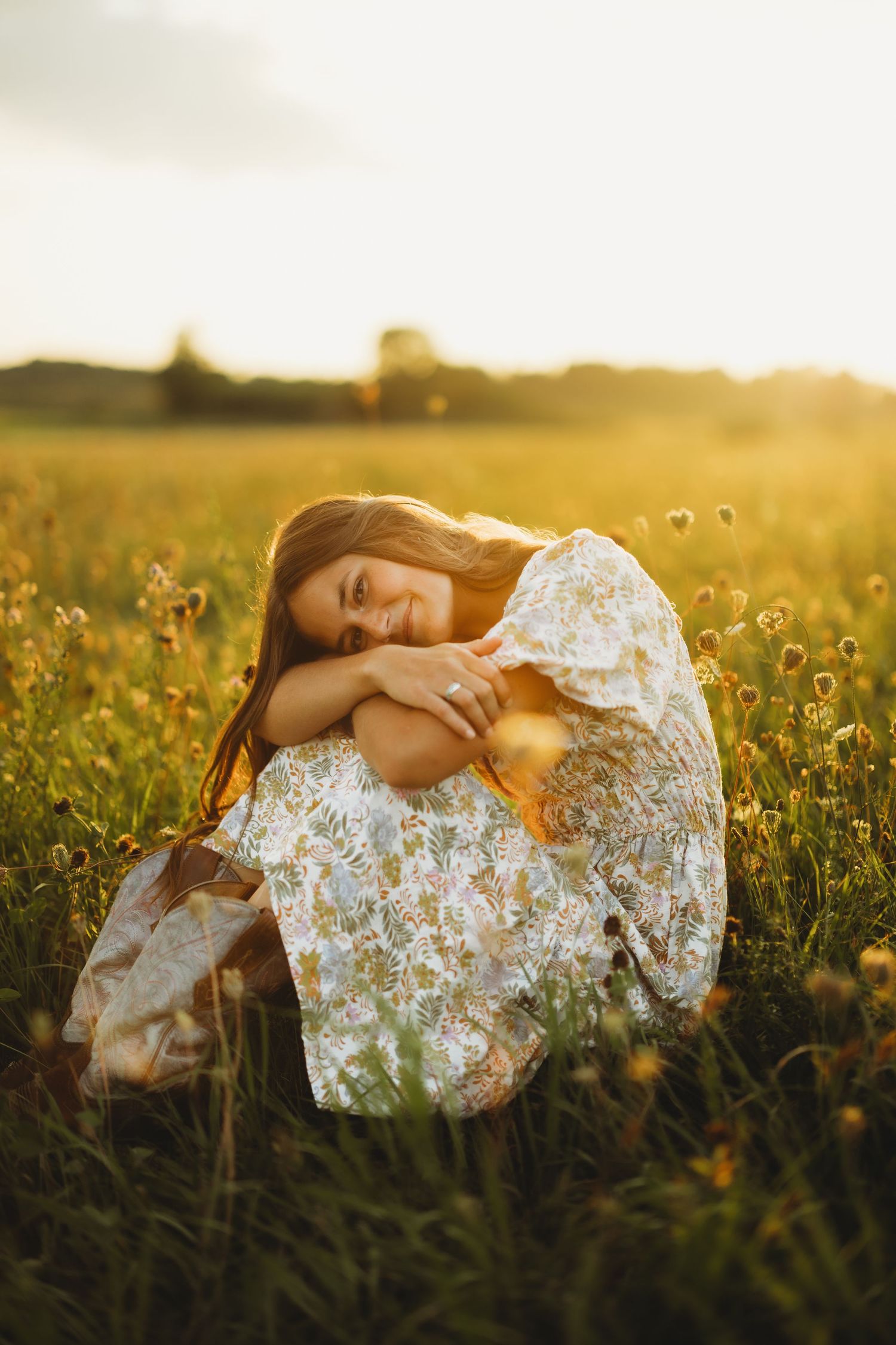 Smiling woman in a floral dress sits hugging her knees in a golden wildflower meadow at sunset.