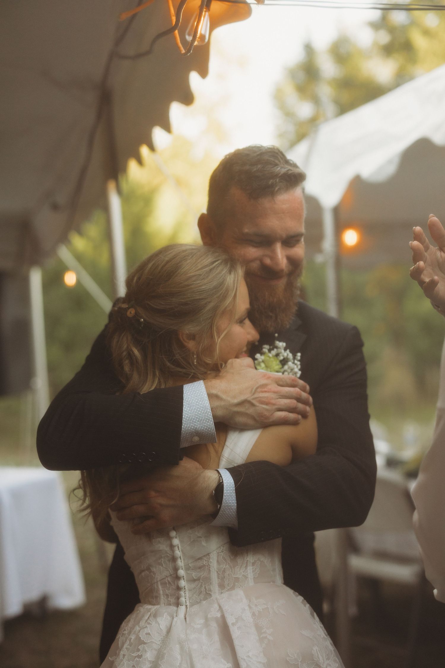 Bearded man in a dark suit embraces a bride in a white lace wedding dress during an outdoor tent reception with warm string lights.