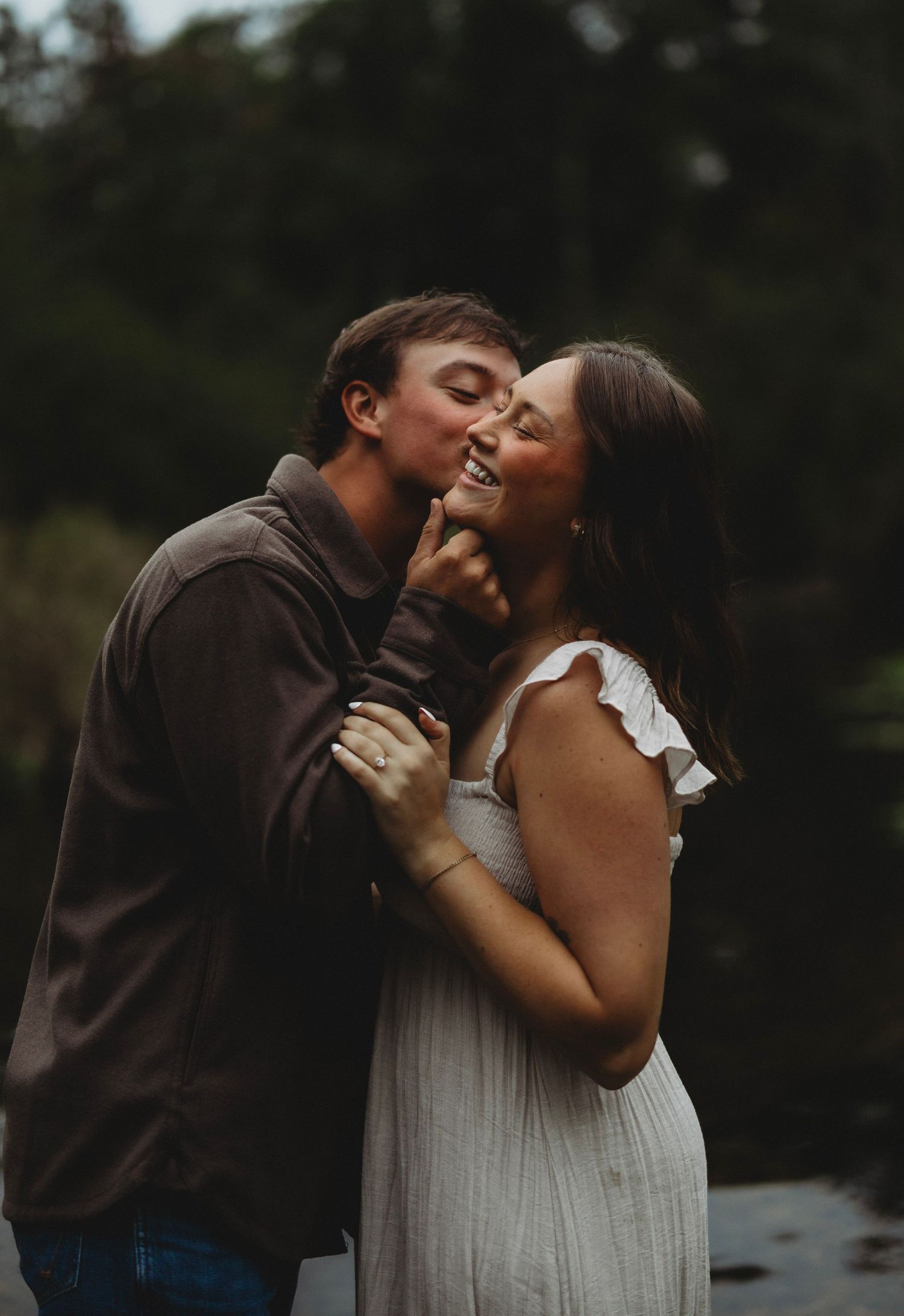 A man in a brown jacket kisses the cheek of a smiling woman in a white sundress during an outdoor couples photo session near the water.