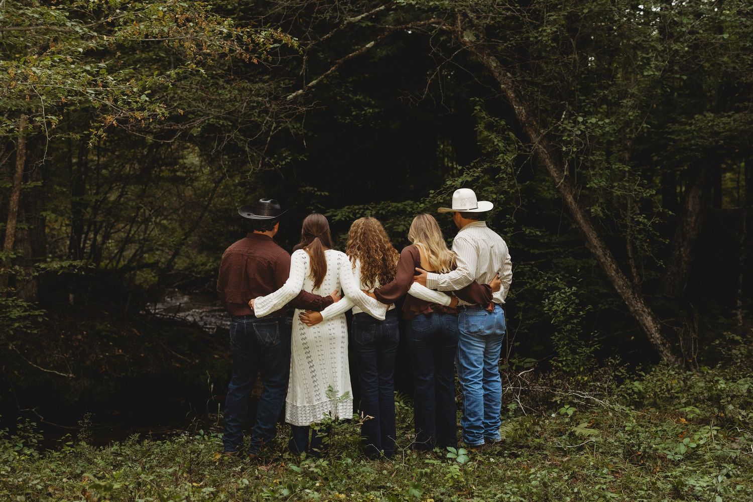 Five people in western attire stand arm in arm with their backs to the camera, facing a dense, shadowy forest.