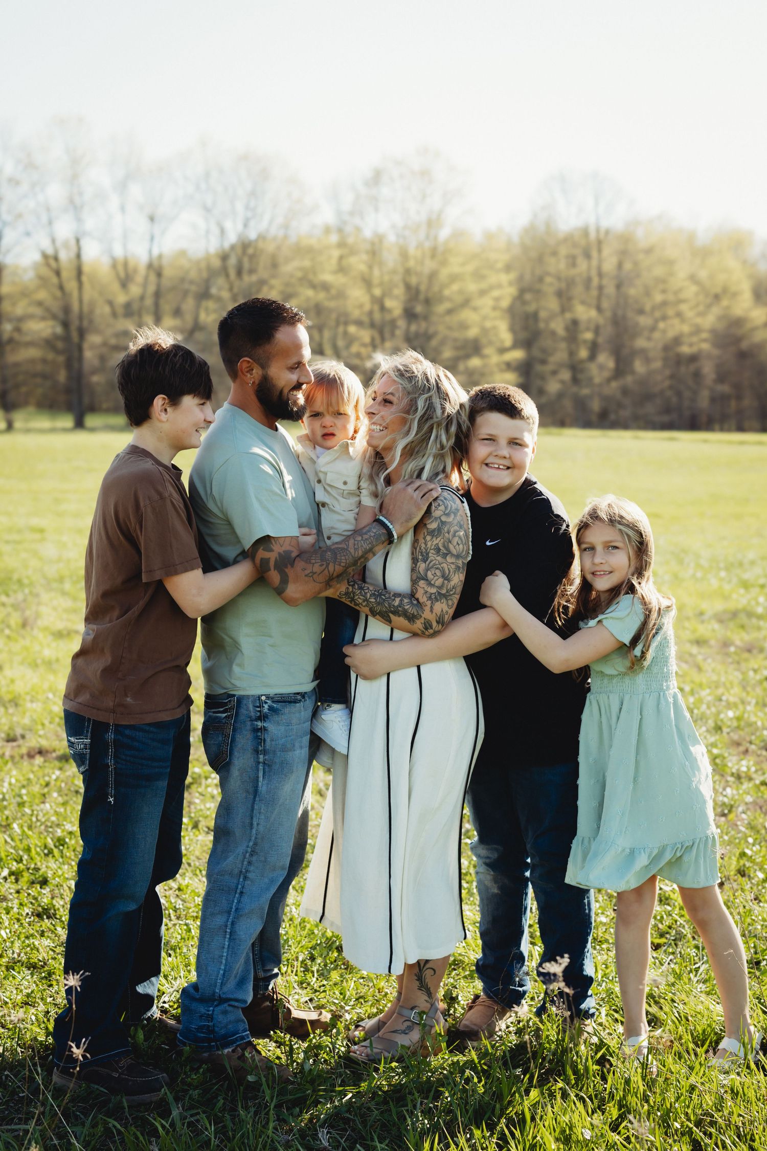 Happy family of six posing together in a sunny green field, parents with four children smiling outdoors.