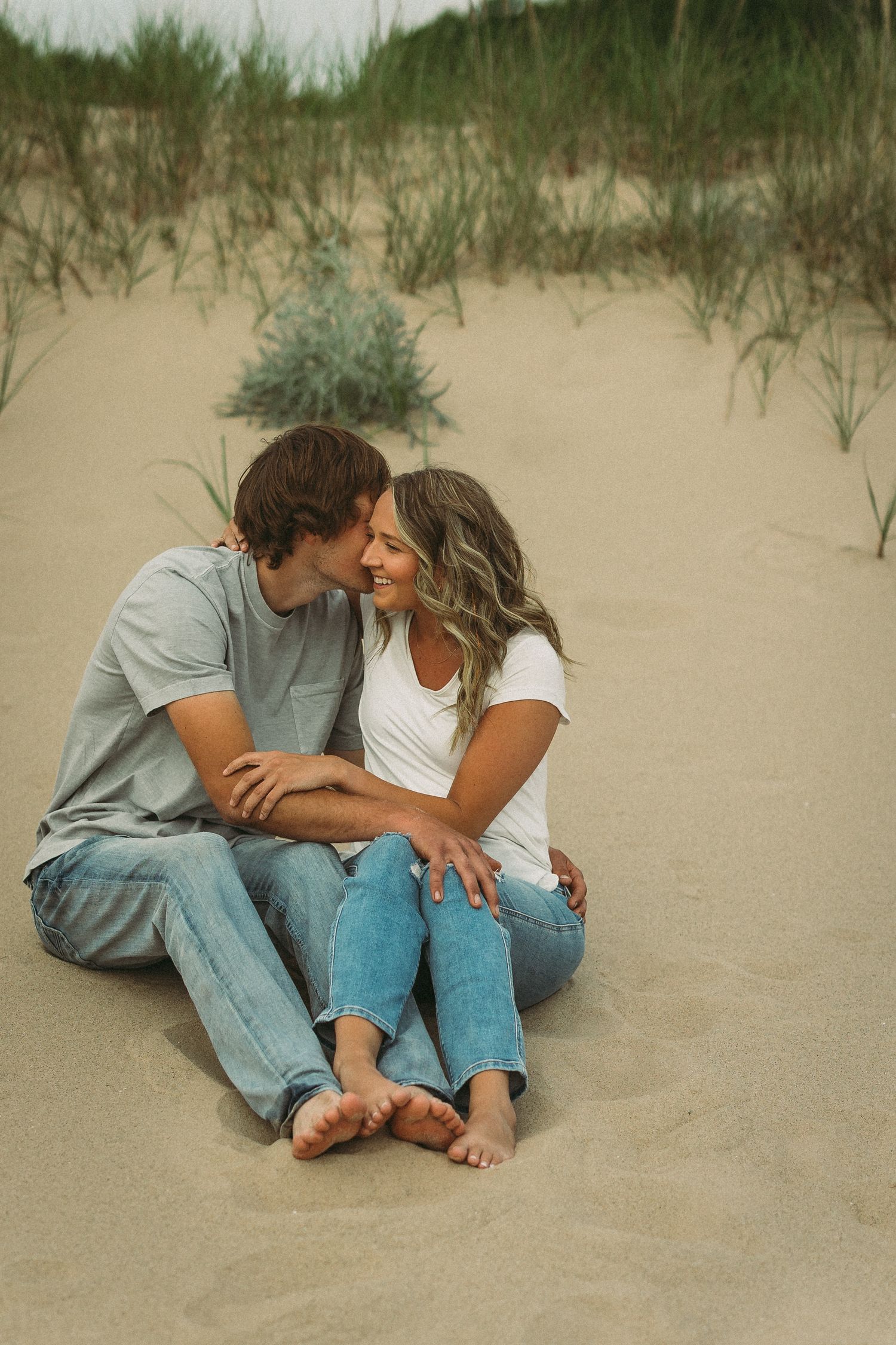 Couple sharing tender moments while sitting in the sand near beach grass.