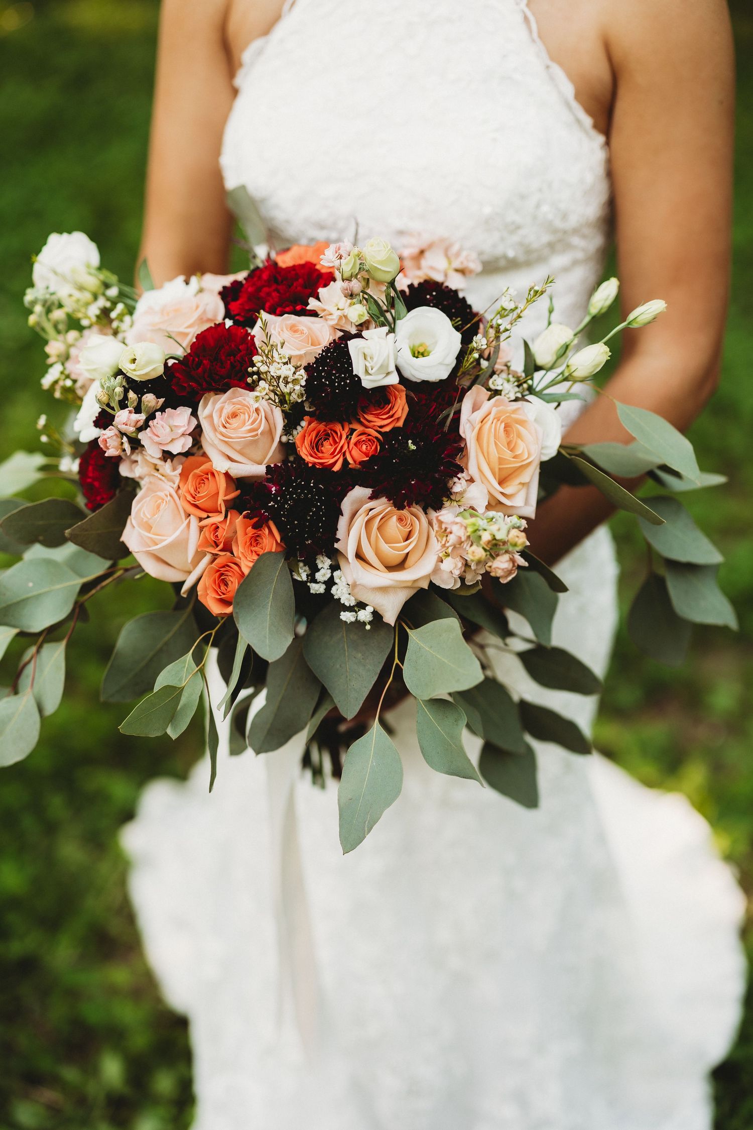 Beautiful wedding bouquet featuring coral roses, deep red flowers, and eucalyptus leaves against a green outdoor backdrop.