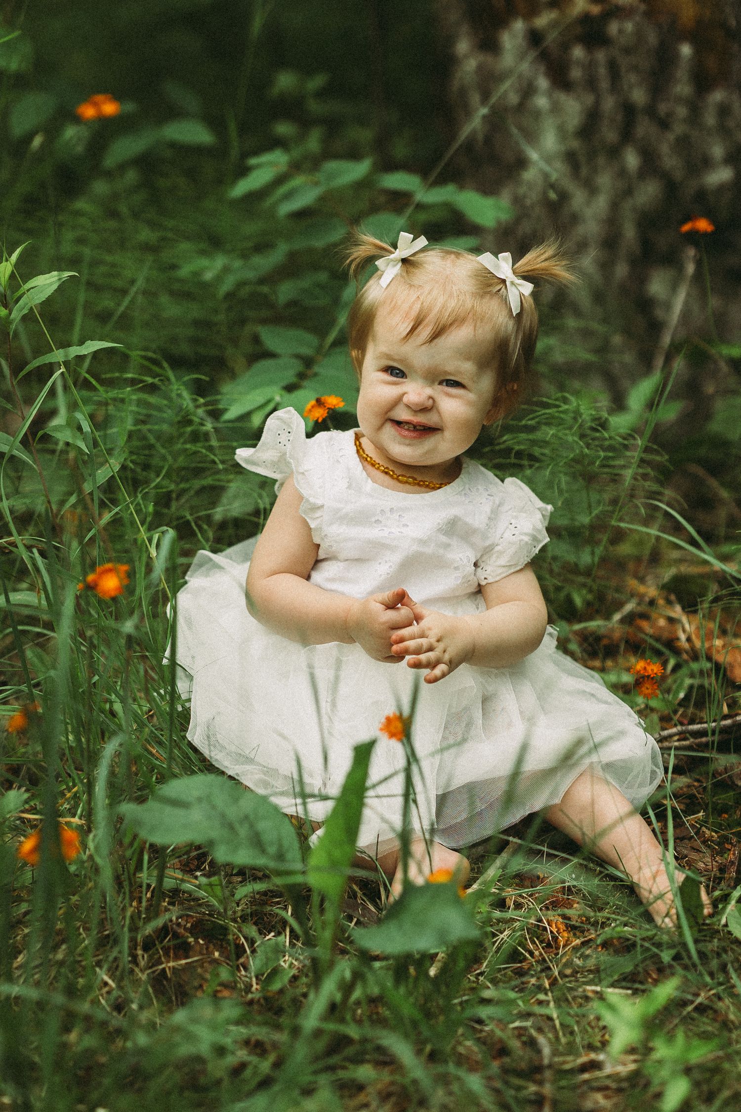 A small child in a white dress sits among green foliage and orange flowers in a garden setting.