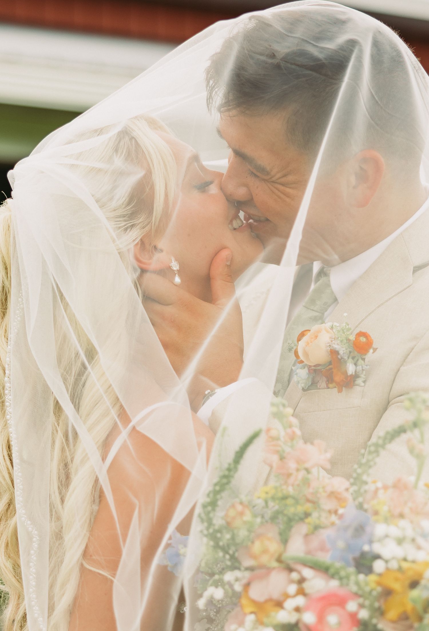 A bride wearing a delicate white veil looks down at colorful floral bouquet in a soft, ethereal wedding portrait.