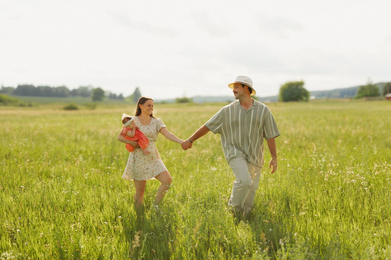 A young family walks hand-in-hand through a sunny green meadow, with a woman carrying a baby and a man in a striped shirt.