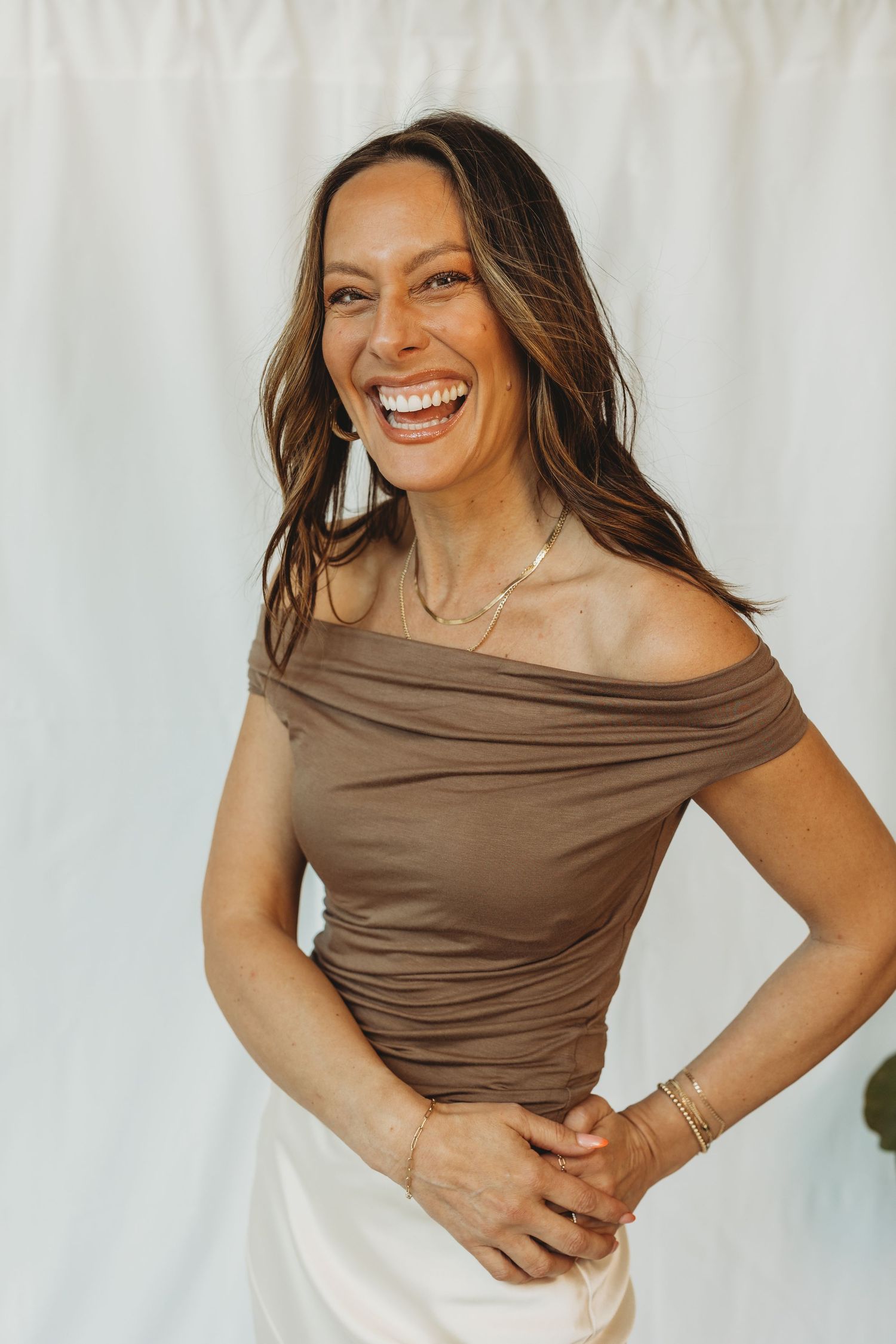 Woman in brown off-shoulder top laughing joyfully, wearing gold jewelry and white skirt against white backdrop.