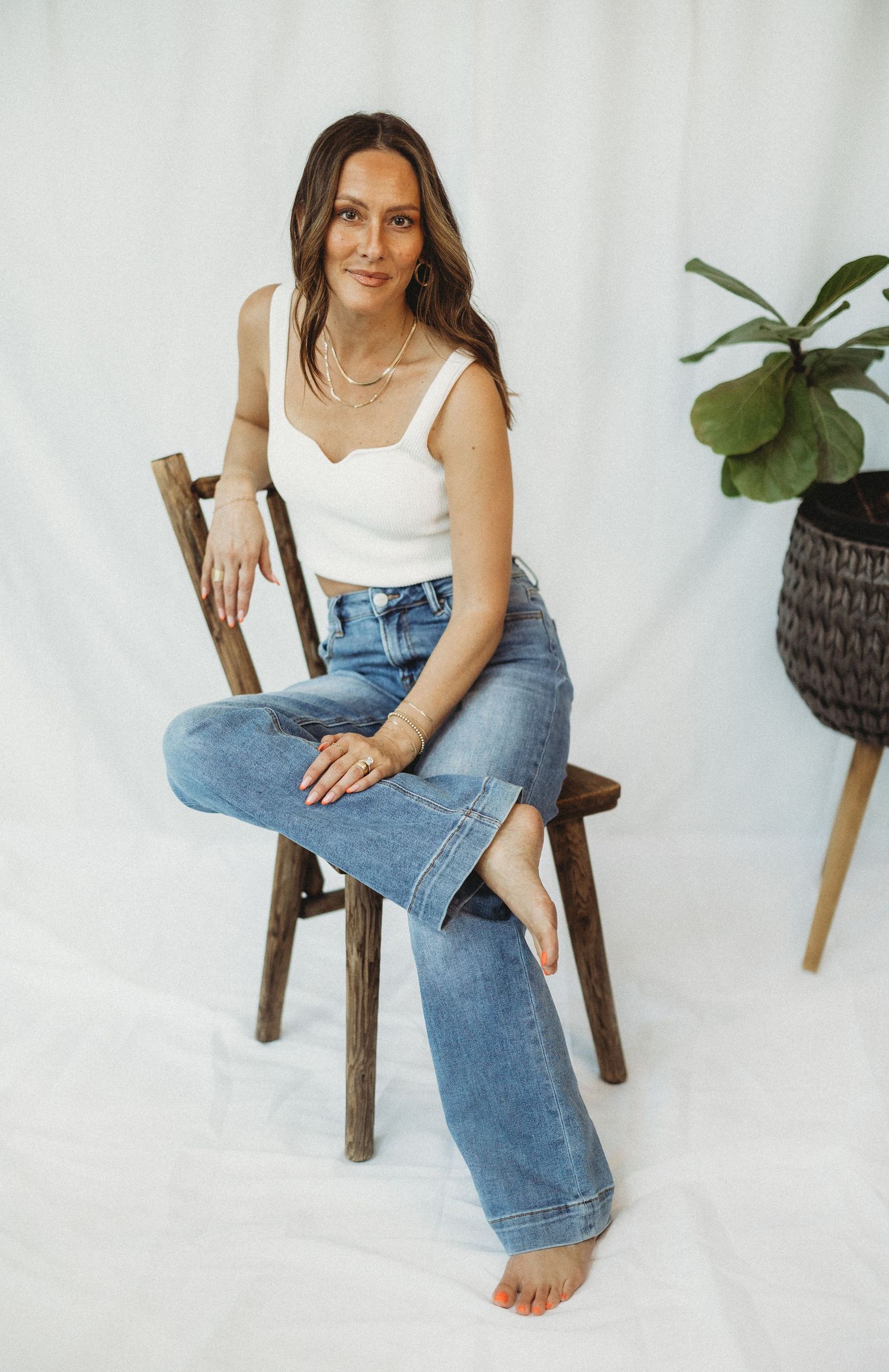Woman sitting on wooden chair wearing white crop top and wide-leg jeans, posed against white backdrop.