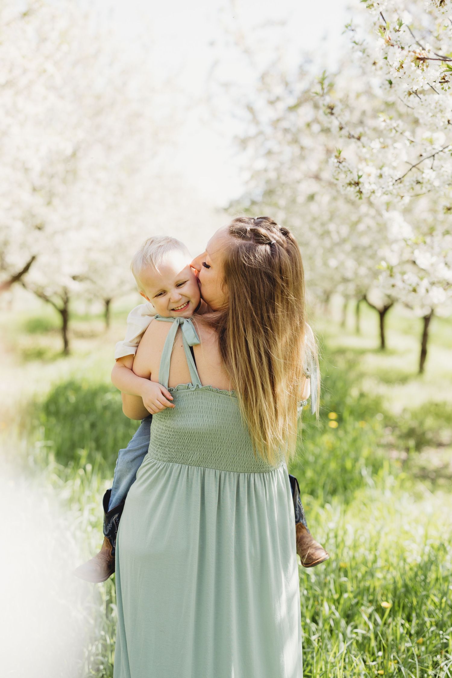 A mother in a sage green dress kisses her smiling toddler among blooming white trees in a spring orchard.