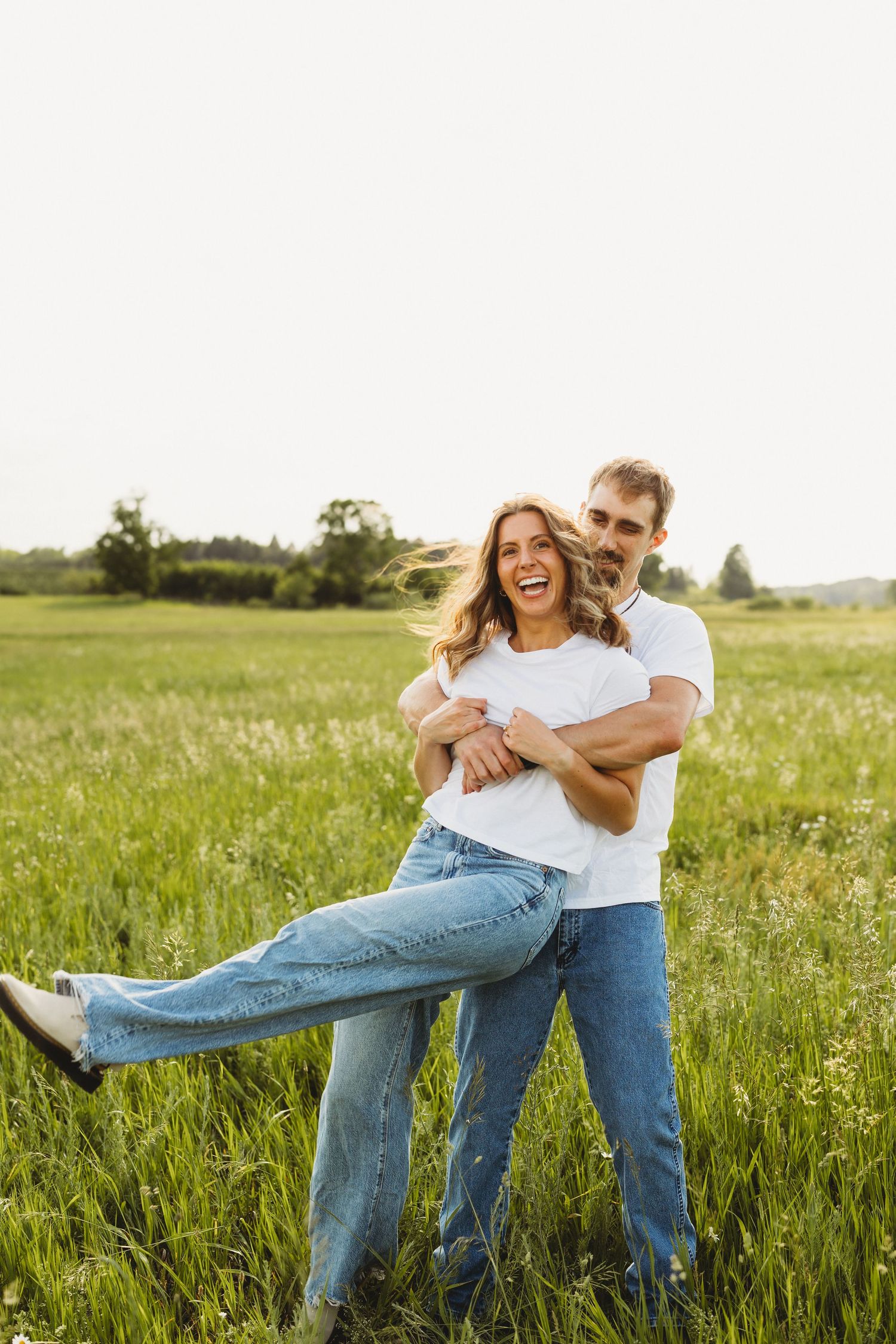 Happy couple laughing and embracing in a sunny green field, both wearing white t-shirts and jeans.