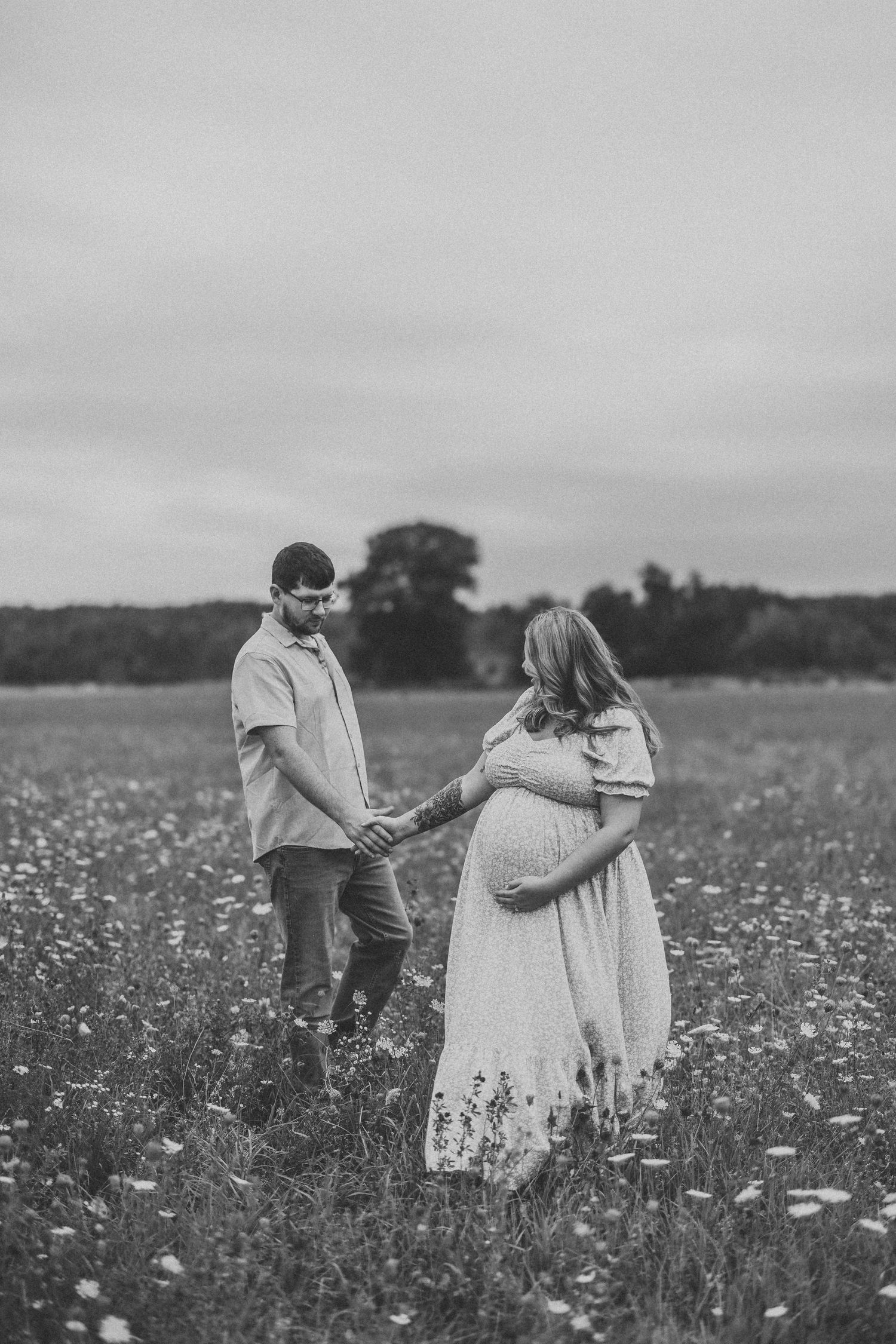 Black and white maternity photo of couple holding hands in a wildflower field, woman cradling baby bump.
