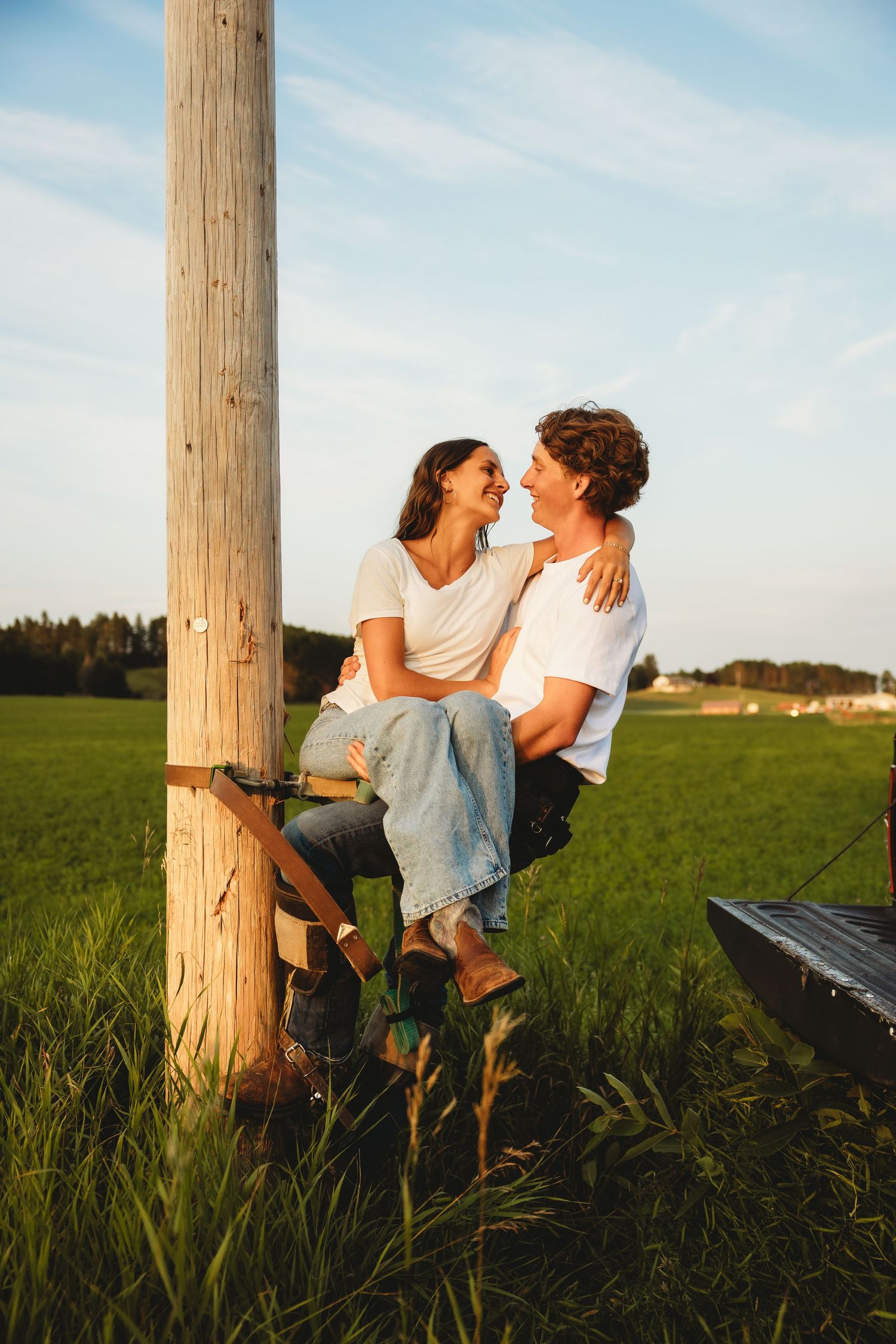 Happy couple in white shirts embrace and smile at each other leaning against a wooden post in a green field.