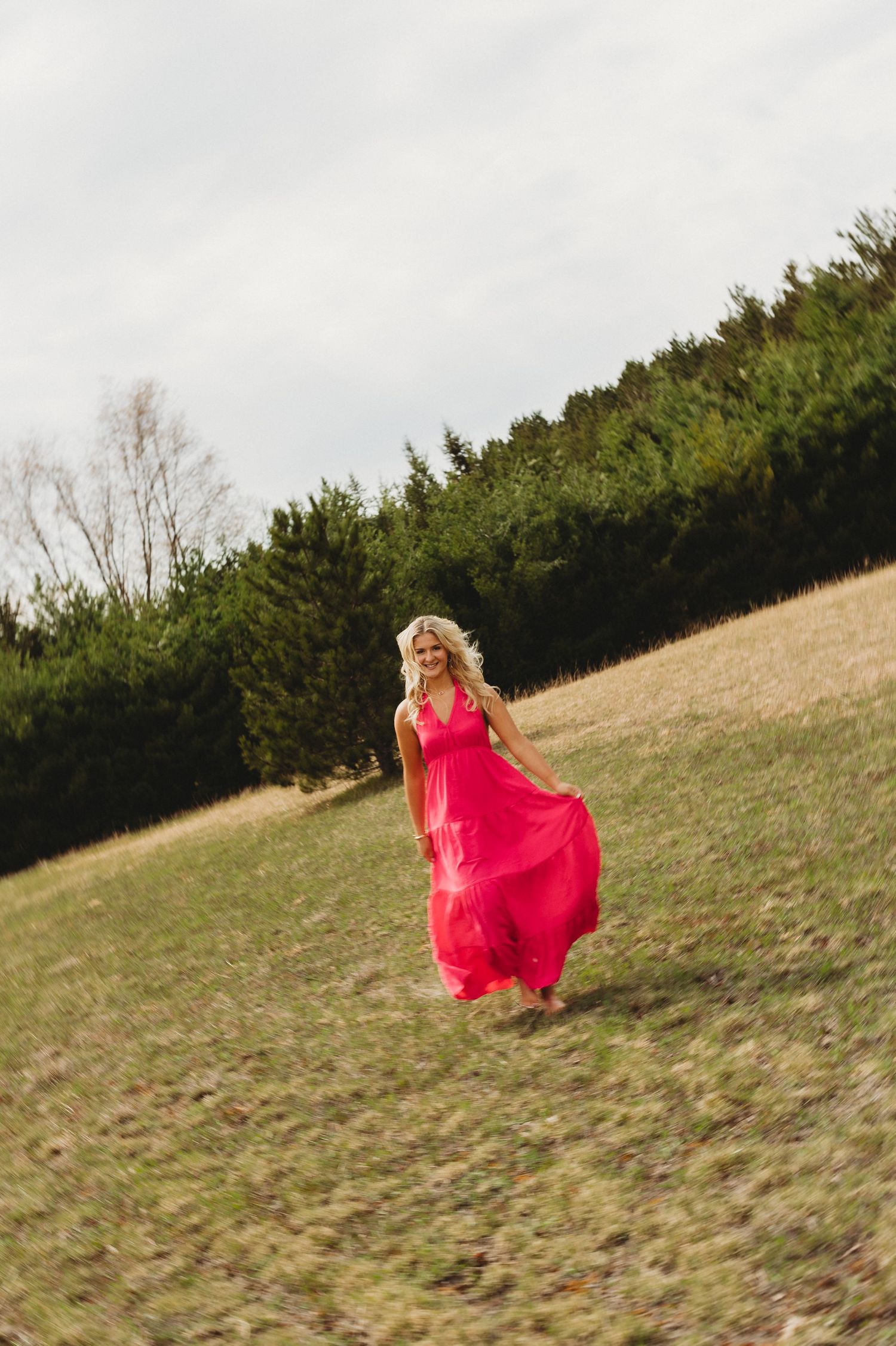 Blonde woman in a vibrant pink maxi dress walking through a grassy field with evergreen trees behind her.