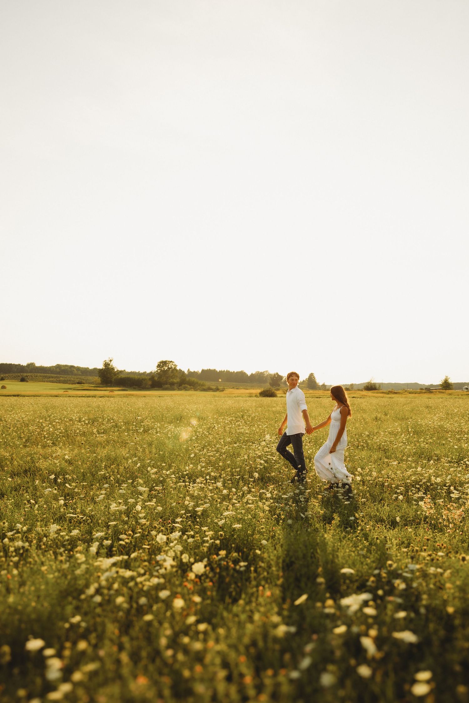 Couple holding hands walking through a sunny wildflower meadow, woman in white dress, man in white shirt.