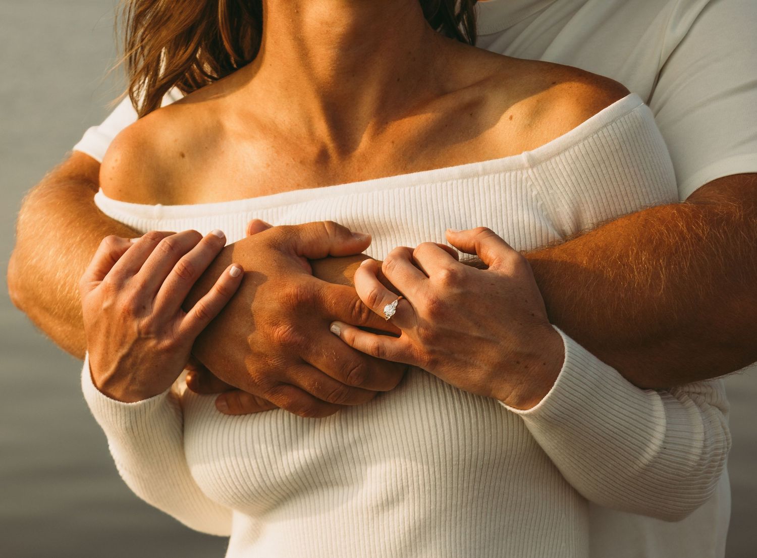 Couple embracing tenderly, man wrapping arms around woman in white off-shoulder sweater, wearing engagement ring.