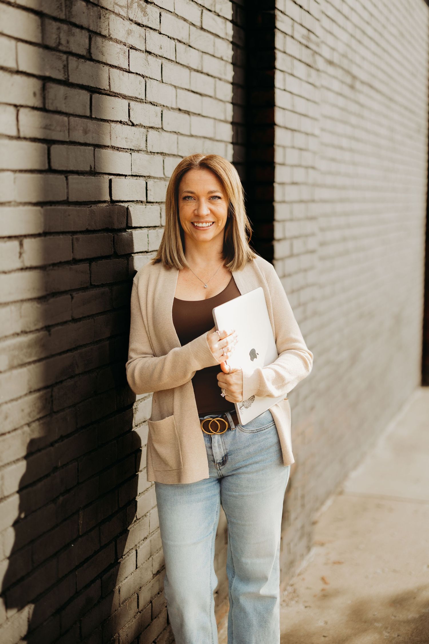 Smiling woman in beige cardigan holding a MacBook laptop, leaning against a dark brick wall in jeans.