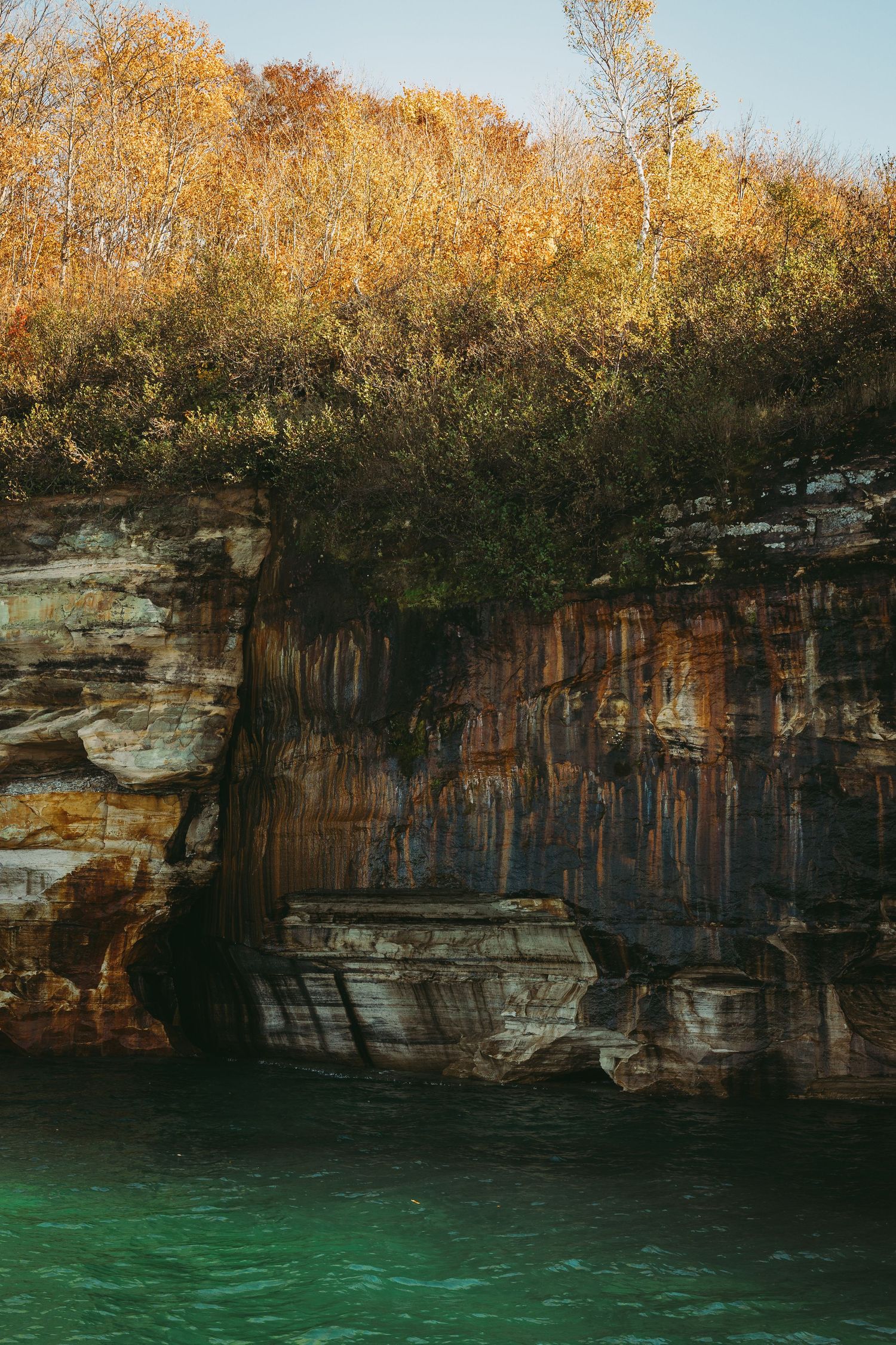 Rugged sandstone cliffs reflect in emerald waters below autumn trees.